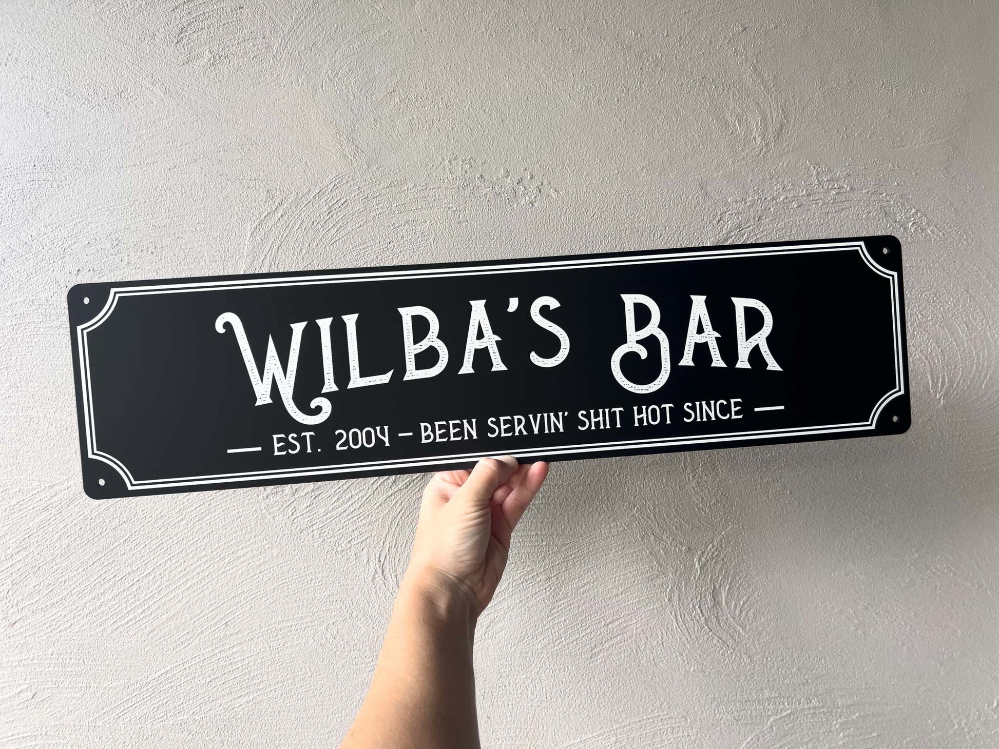Hand holding a black sign with 'Wilba's Bar' text against a light gray wall.