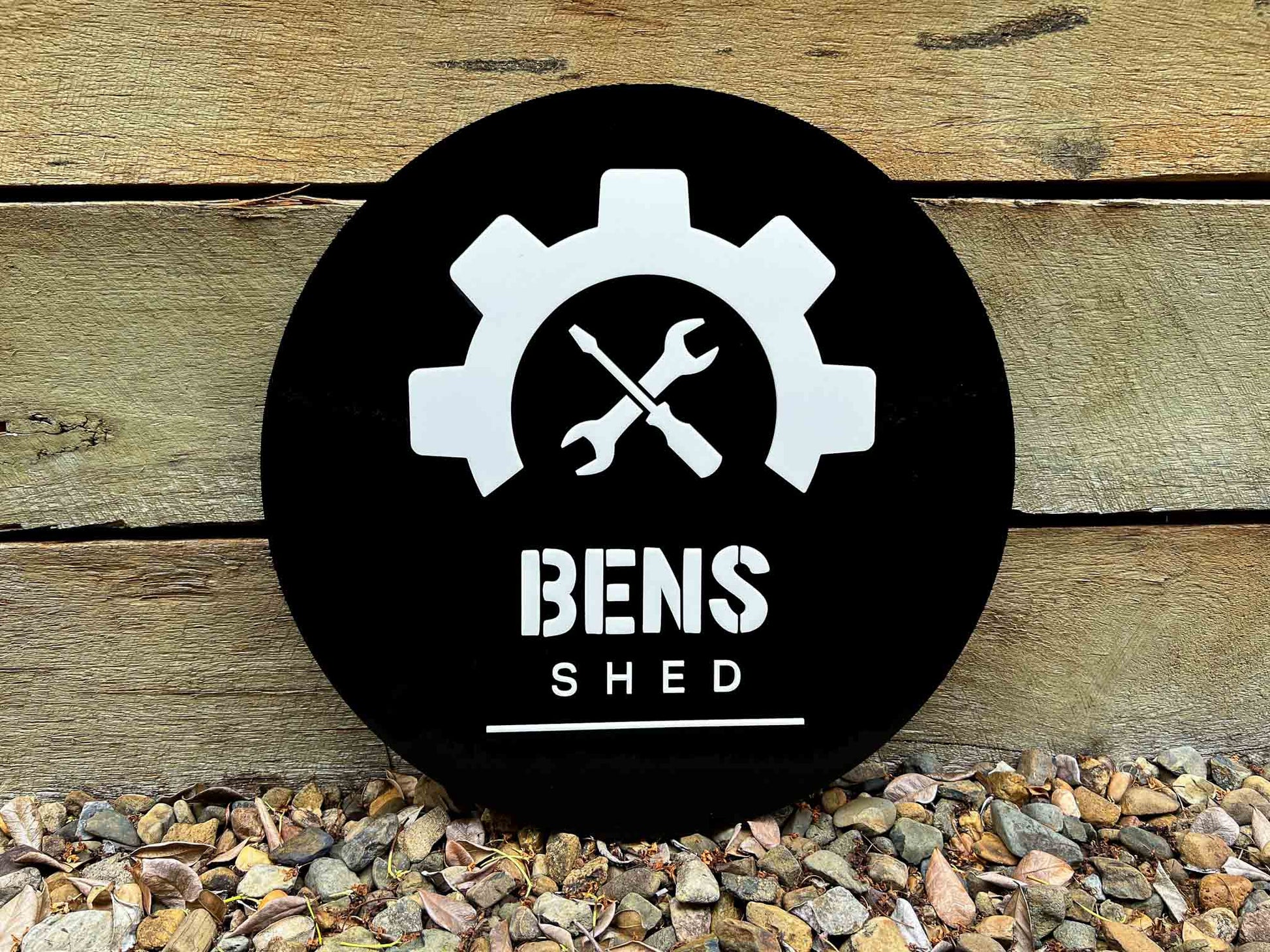 Black circular sign with white gear and wrench design, and 'BENS SHED' text on a wooden background.