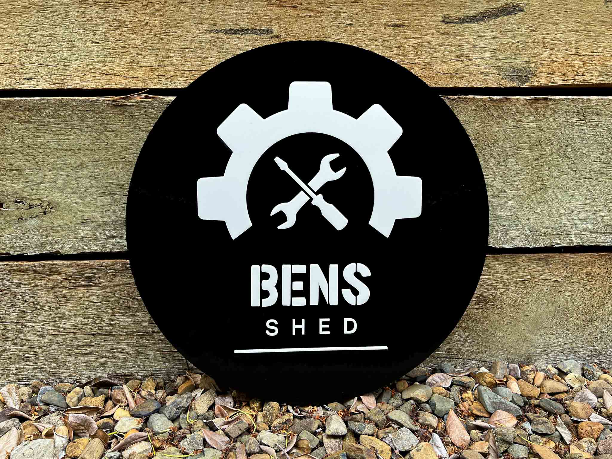 Black circular sign with white gear and wrench design, and 'BENS SHED' text on a wooden background.