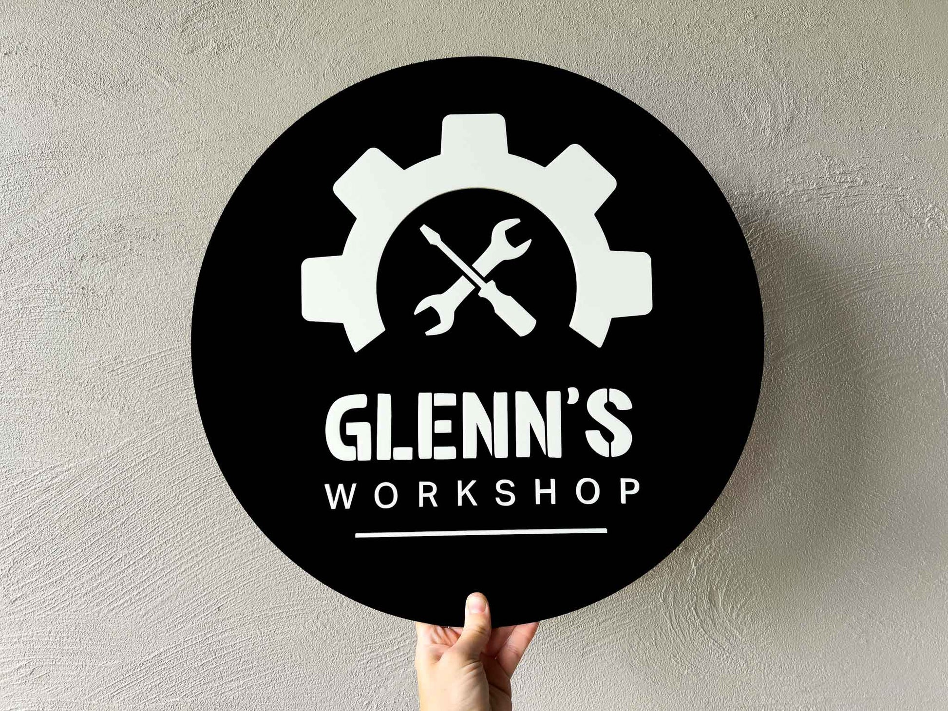 3D acrylic personalised round black sign with white gear and wrench design, and 'Glenn's Workshop' text on a light gray background.