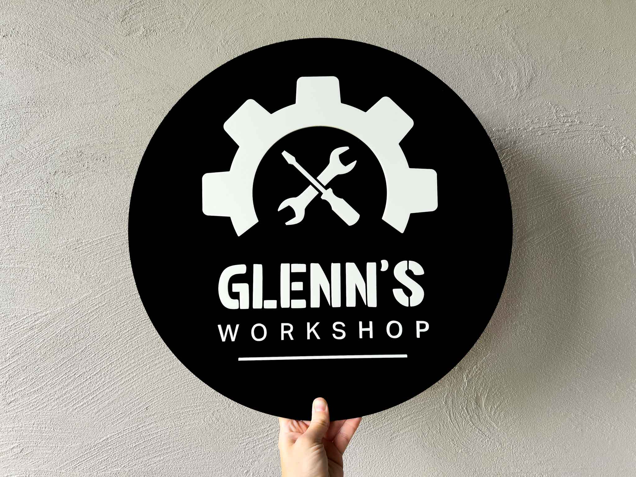 3D acrylic personalised round black sign with white gear and wrench design, and 'Glenn's Workshop' text on a light gray background.