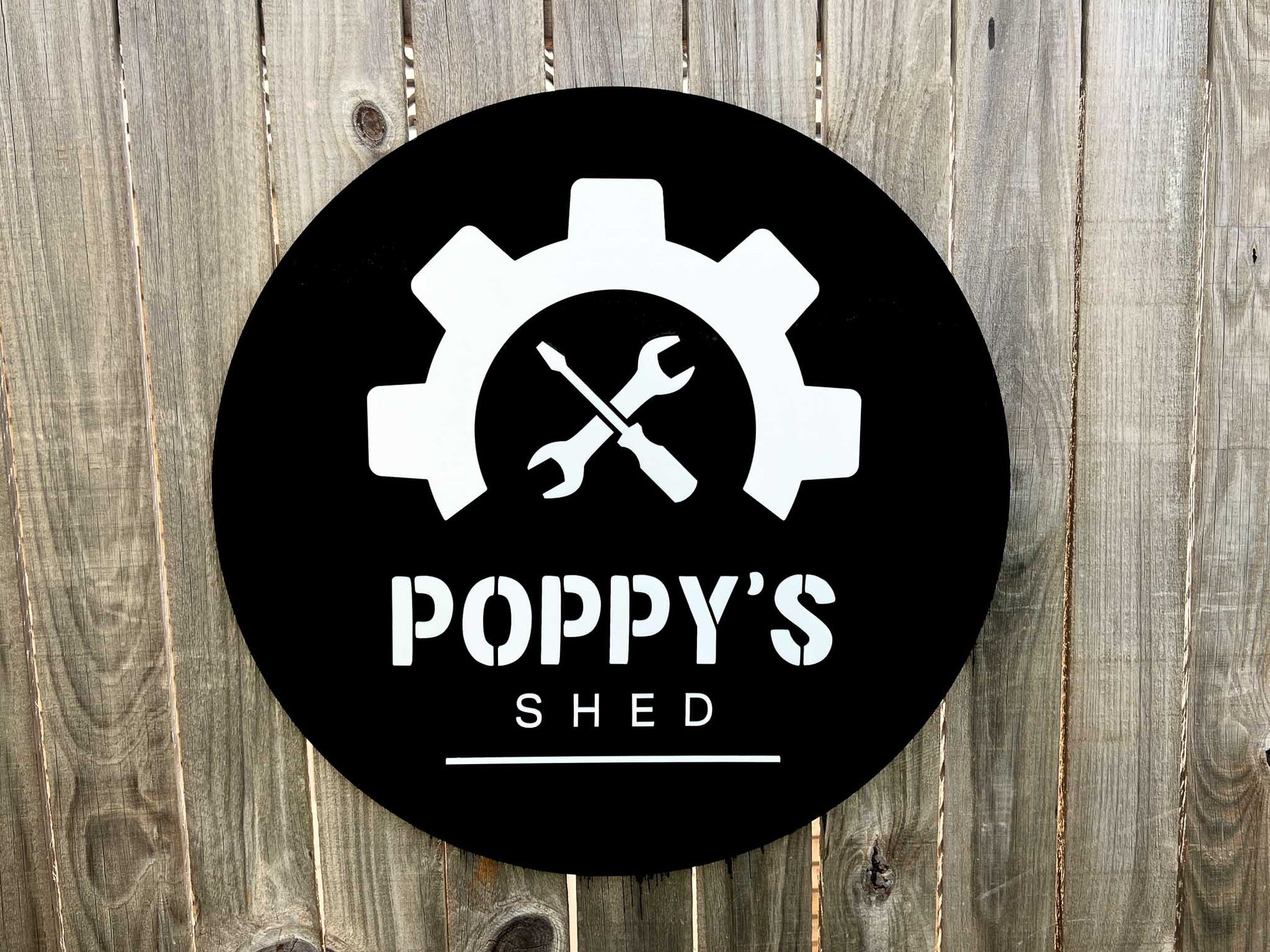Custom made round black sign with 'Poppy's Shed' logo featuring gears and crossed wrenches on a wooden background.