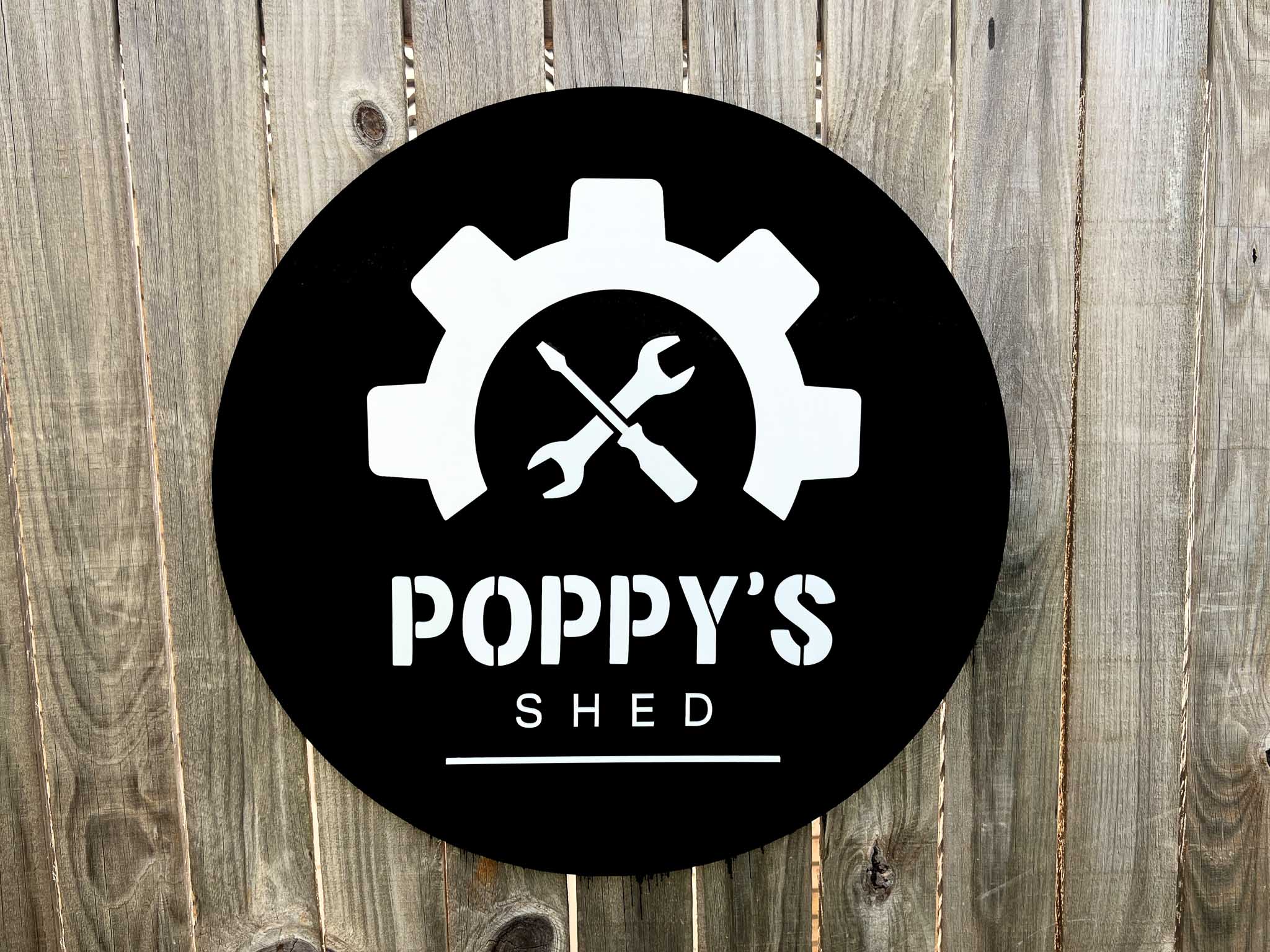 Custom made round black sign with 'Poppy's Shed' logo featuring gears and crossed wrenches on a wooden background.