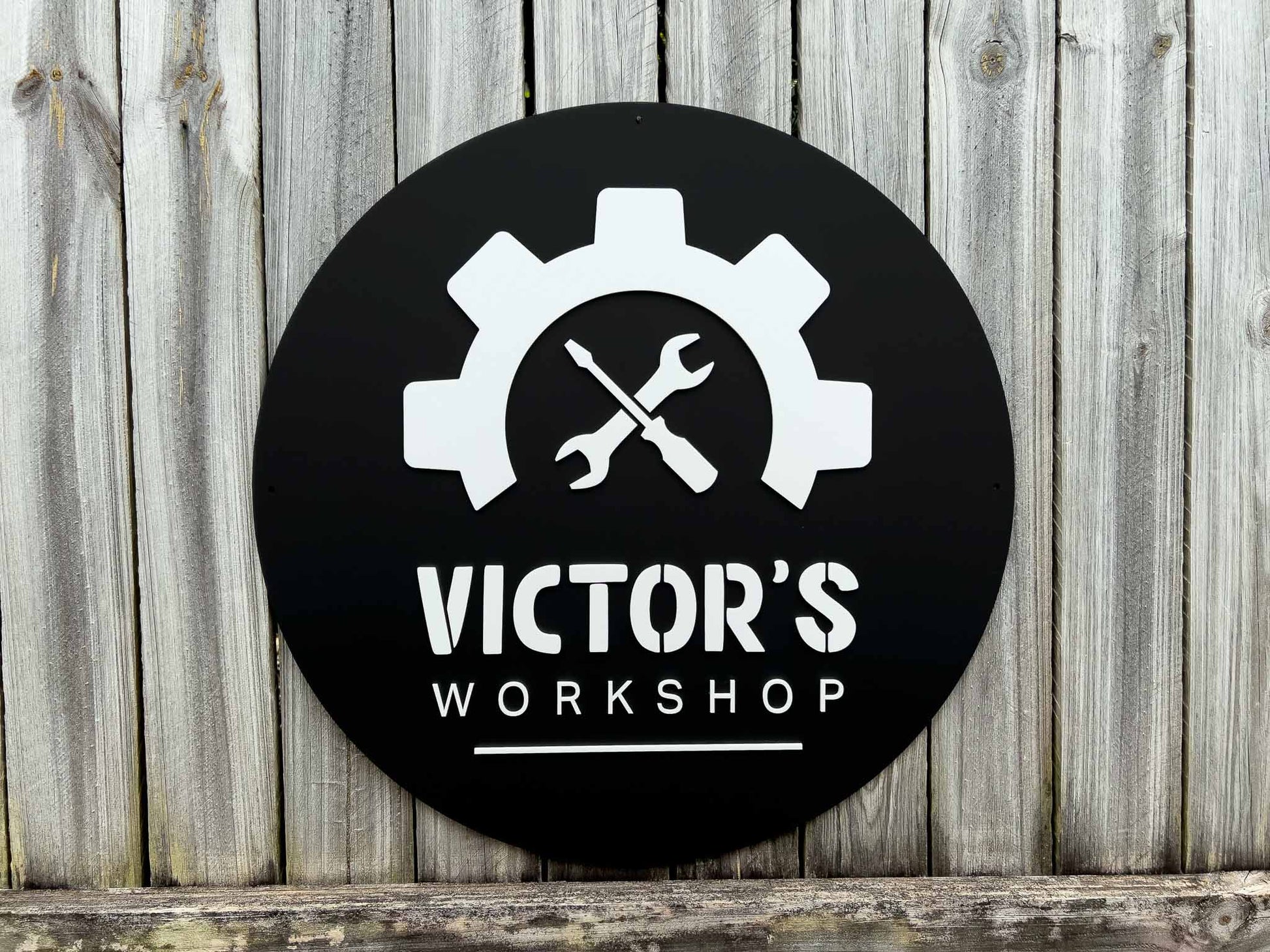 Round black sign with white gear and wrench design, and 'Victor's Workshop' text on a wooden background.
