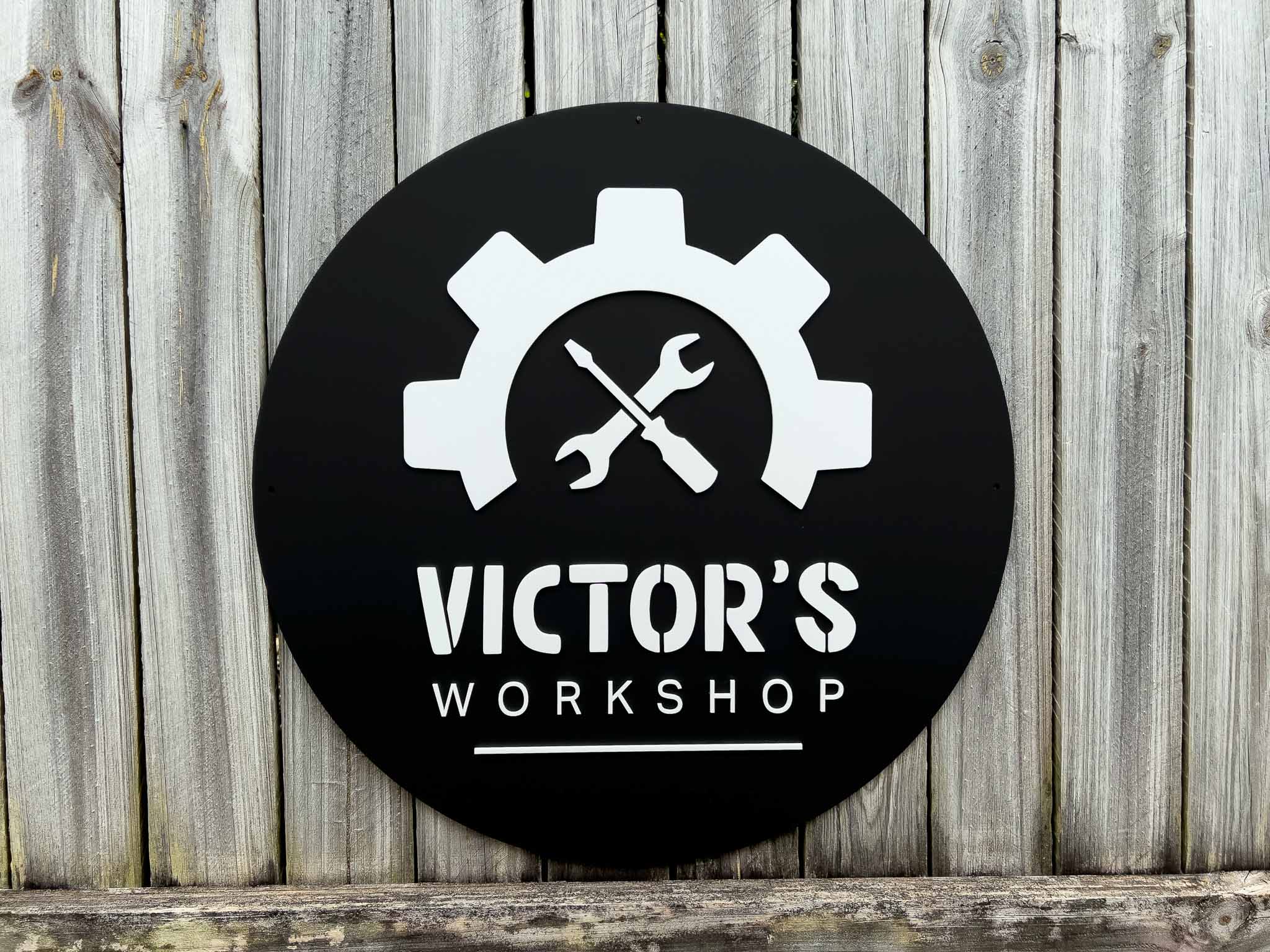 Round black sign with white gear and wrench design, and 'Victor's Workshop' text on a wooden background.