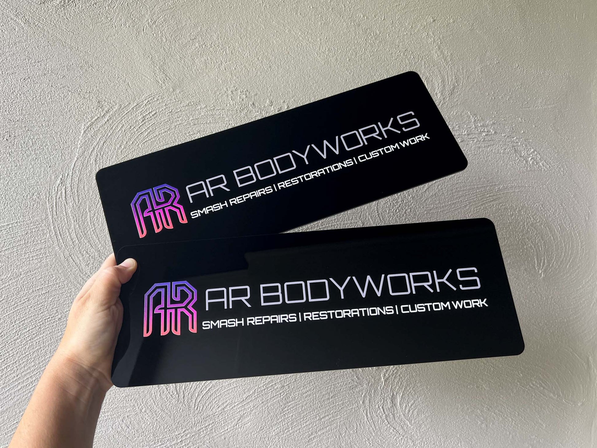 Two black signs with 'AR Bodyworks' logo and text held against a light grey background.