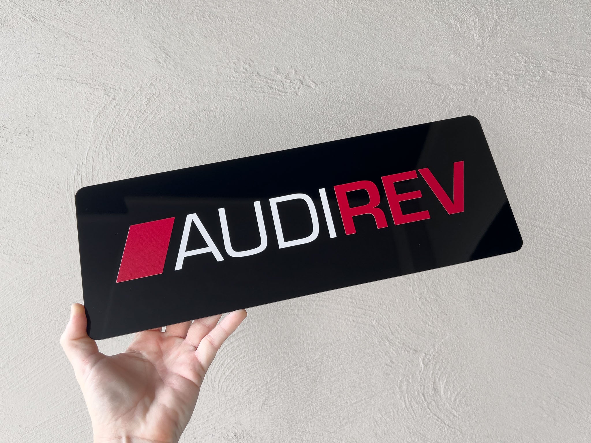 Hand holding a black logo plate sign with an 'AUDIREV' logo against a light grey background