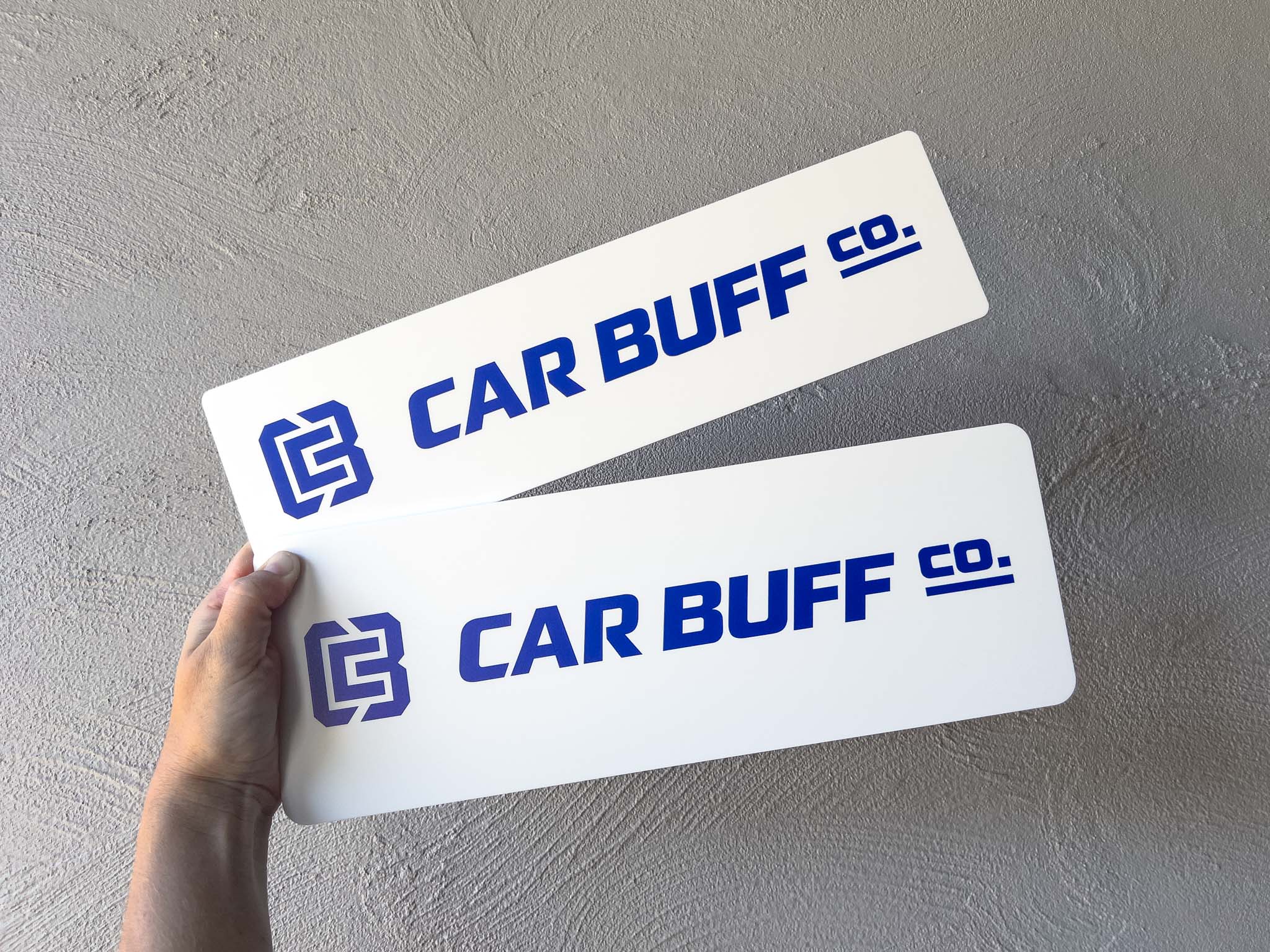 Two 'CAR BUFF CO.' logo cover plates held by a hand on a grey background