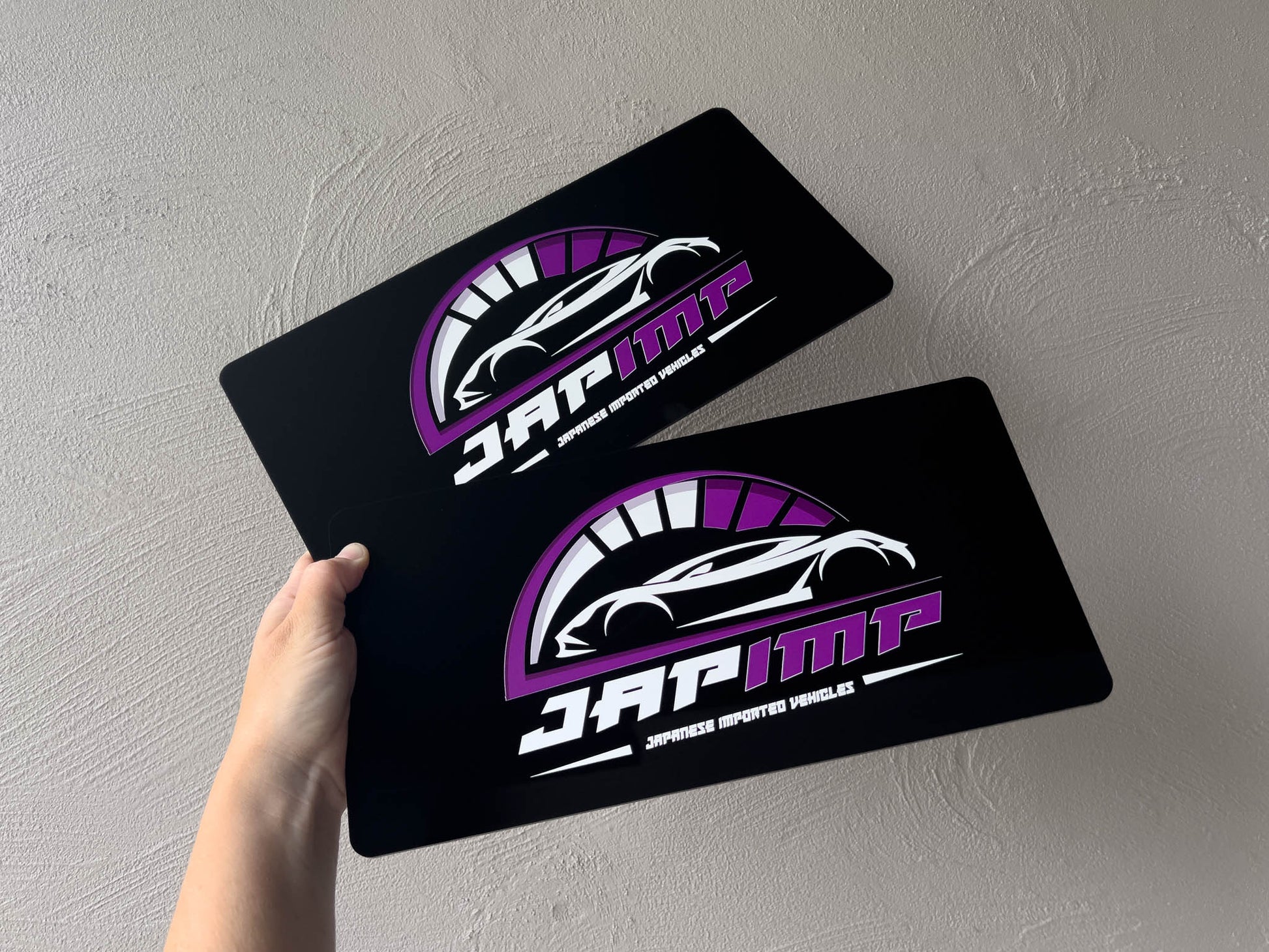 Two black acrylic USA logo plates with a car logo and 'JAP IMP' text held against a grey wall.