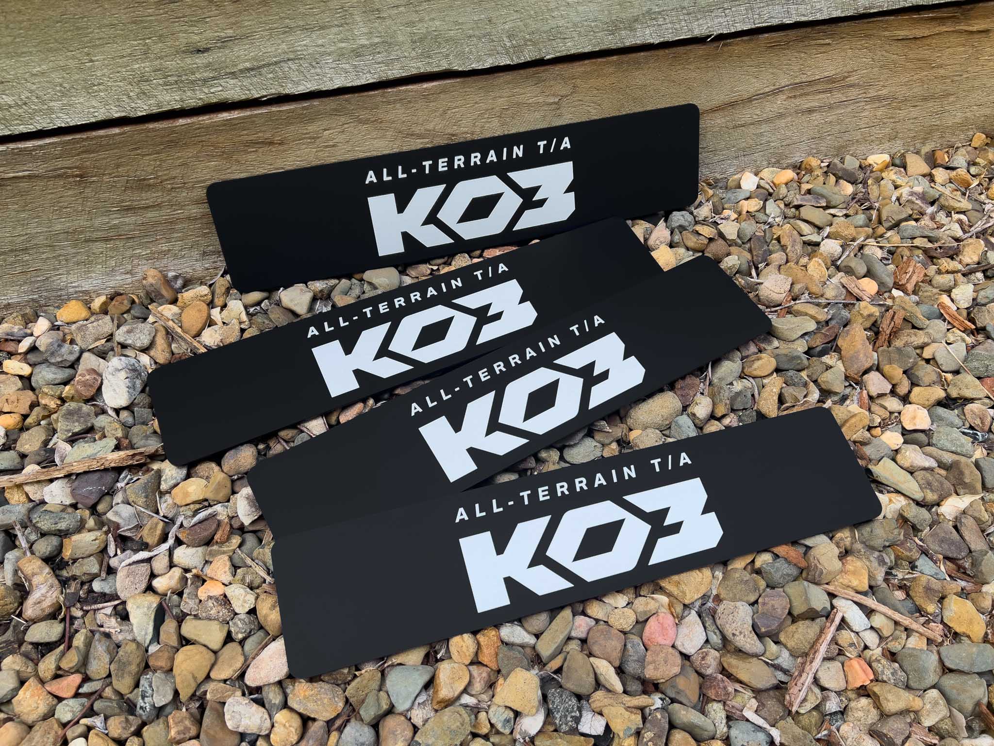 Four black KOZ All-Terrain custom printed logo plates on a gravel surface