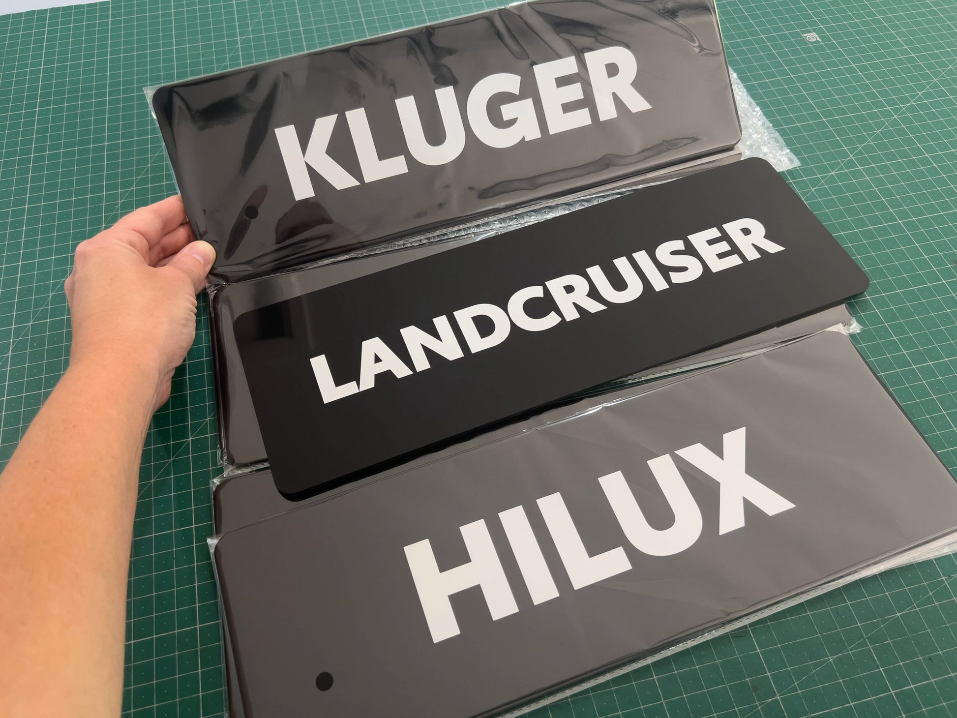 Hand holding a set of car branded logo plates labeled 'Kluger', 'Landcruiser', and 'Hilux' on table.