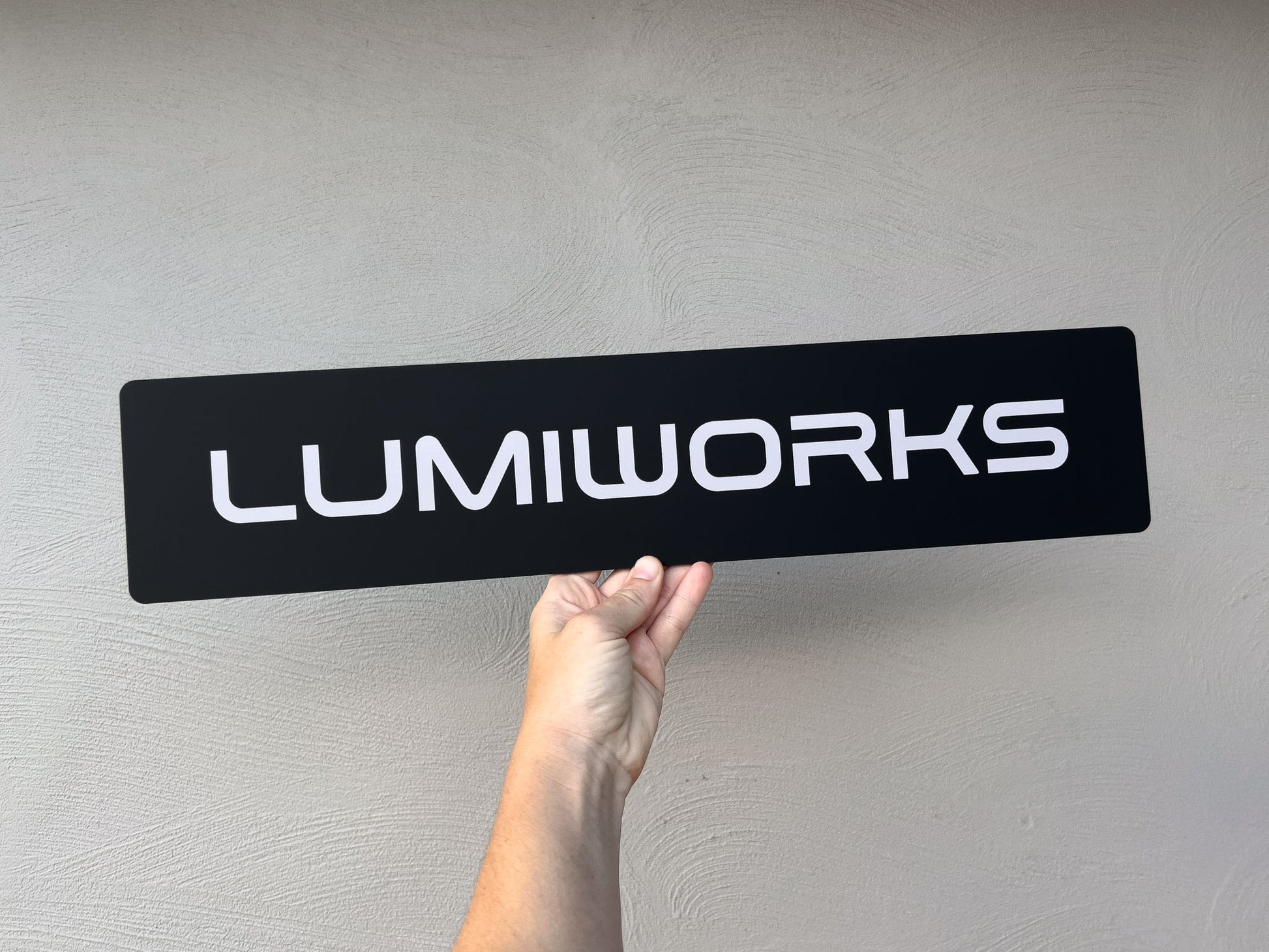 Hand holding a black Euro logo plate sign with 'LUMIWORKS' in white against a grey background