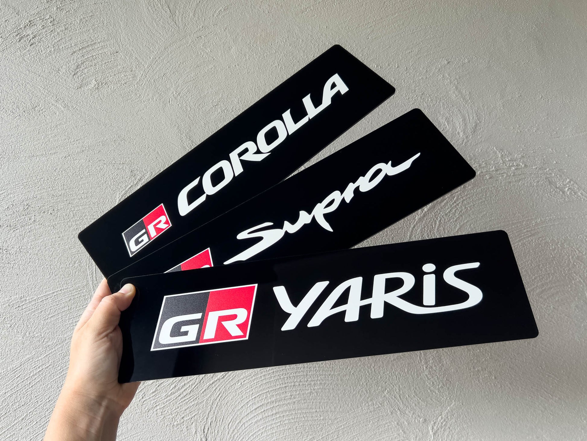 Three acrylic logo plates for Corolla, Supra, and Yaris held by a hand against a light grey background.