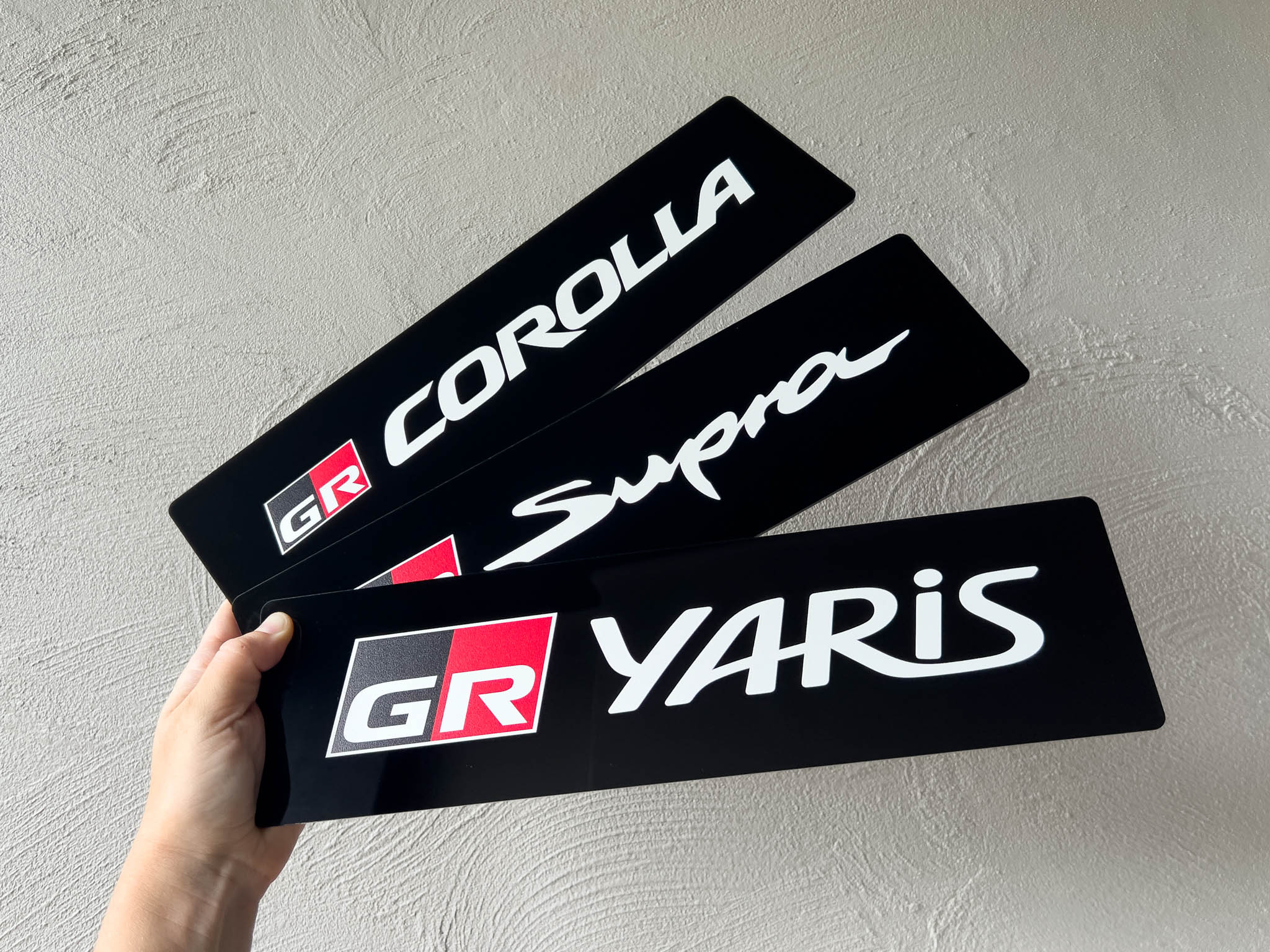 Three acrylic logo plates for Corolla, Supra, and Yaris held by a hand against a light grey background.