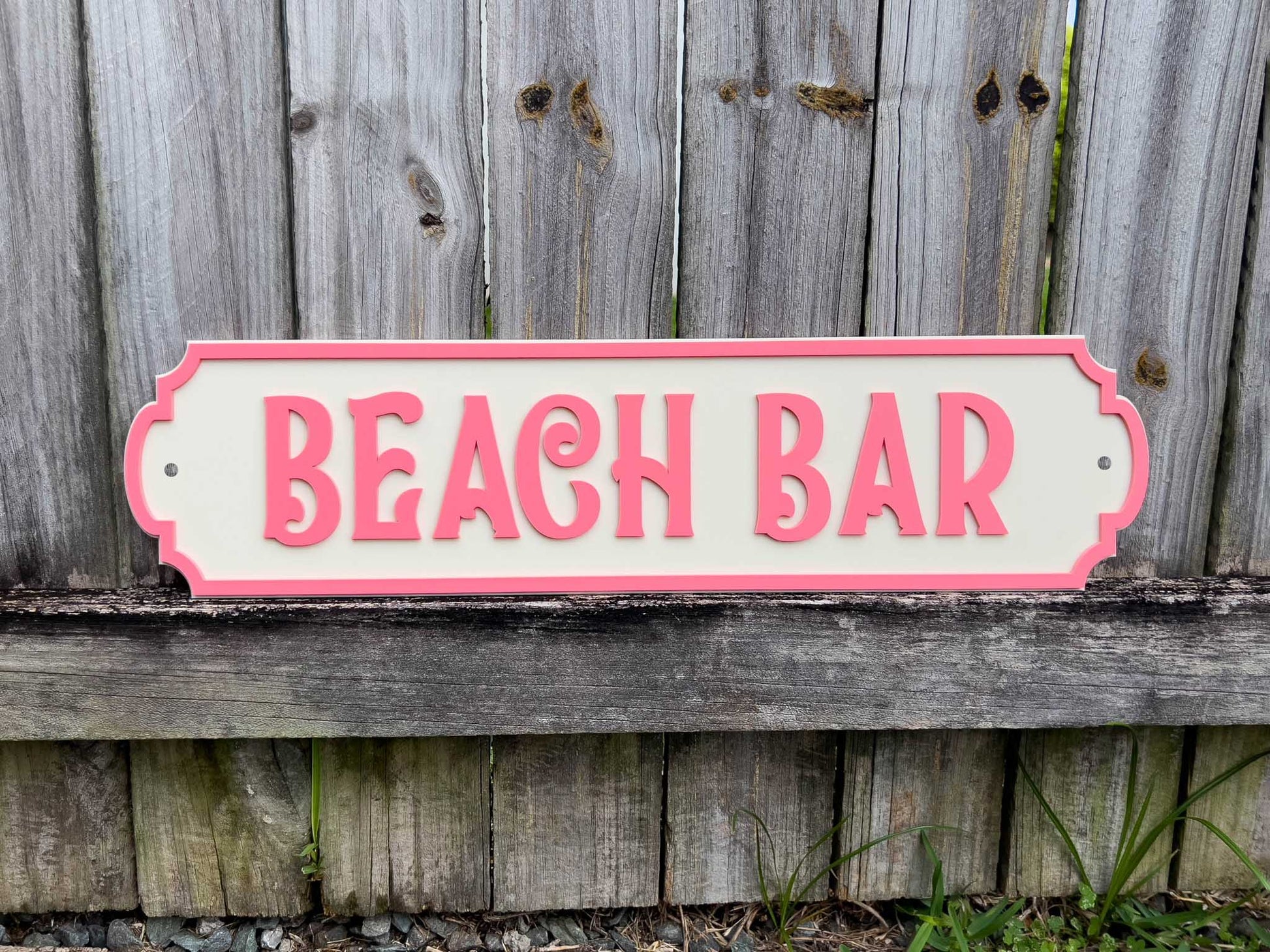 Fresh coastal beach house style sign with 'BEACH BAR' text sitting on a wooden fence.
