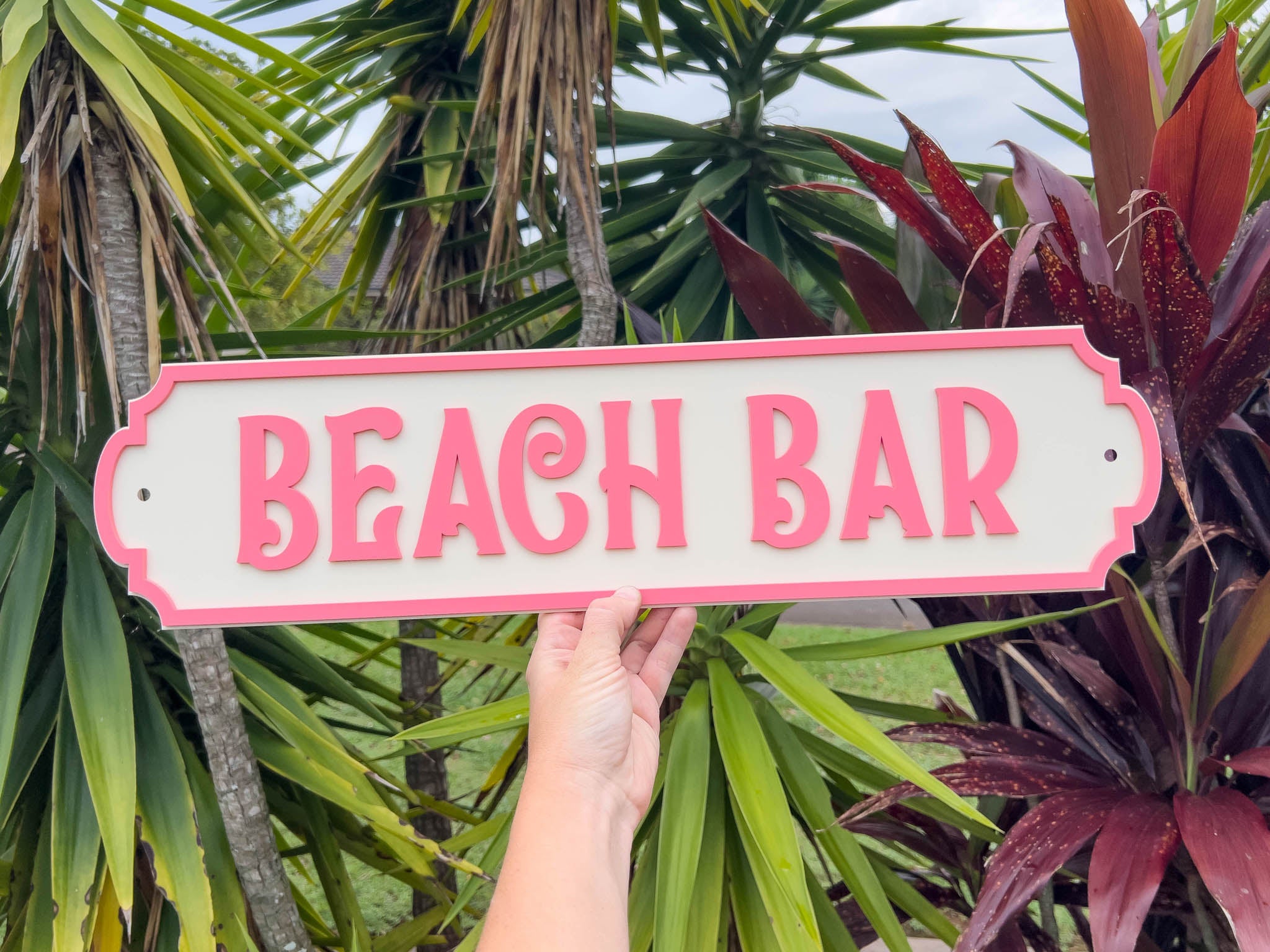Hand holding a 'Beach Bar' sign in front of tropical plants.