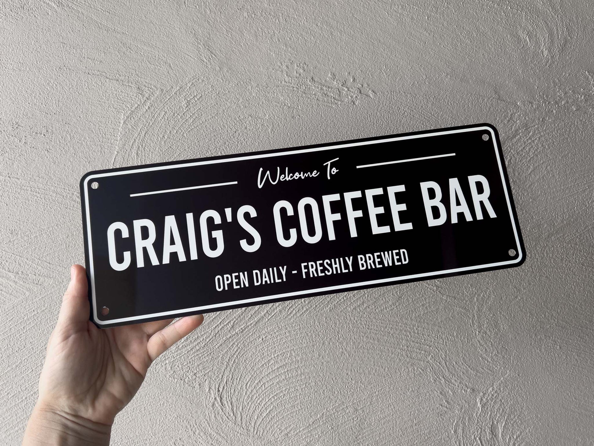 Hand holding a sign that reads 'Welcome to Craig's Coffee Bar Open Daily - Freshly Brewed' against a grey background.