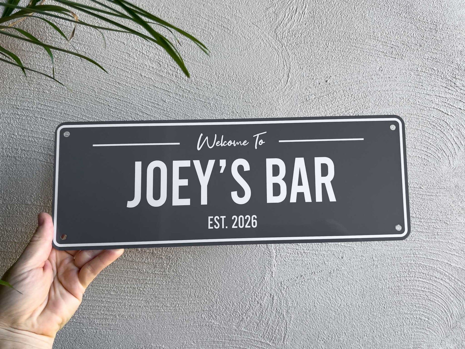 A grey and white custom printed bar sign being held by a hand near a palm tree that says 'Joey's Bar Est. 2026'.
