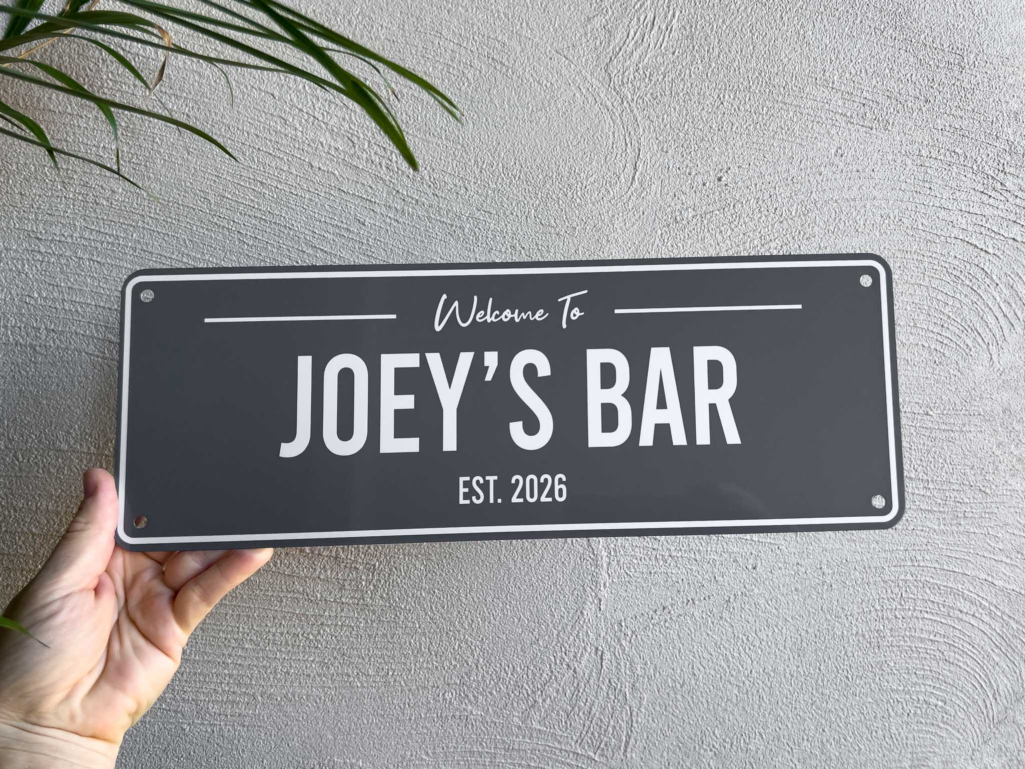 A grey and white custom printed bar sign being held by a hand near a palm tree that says 'Joey's Bar Est. 2026'.