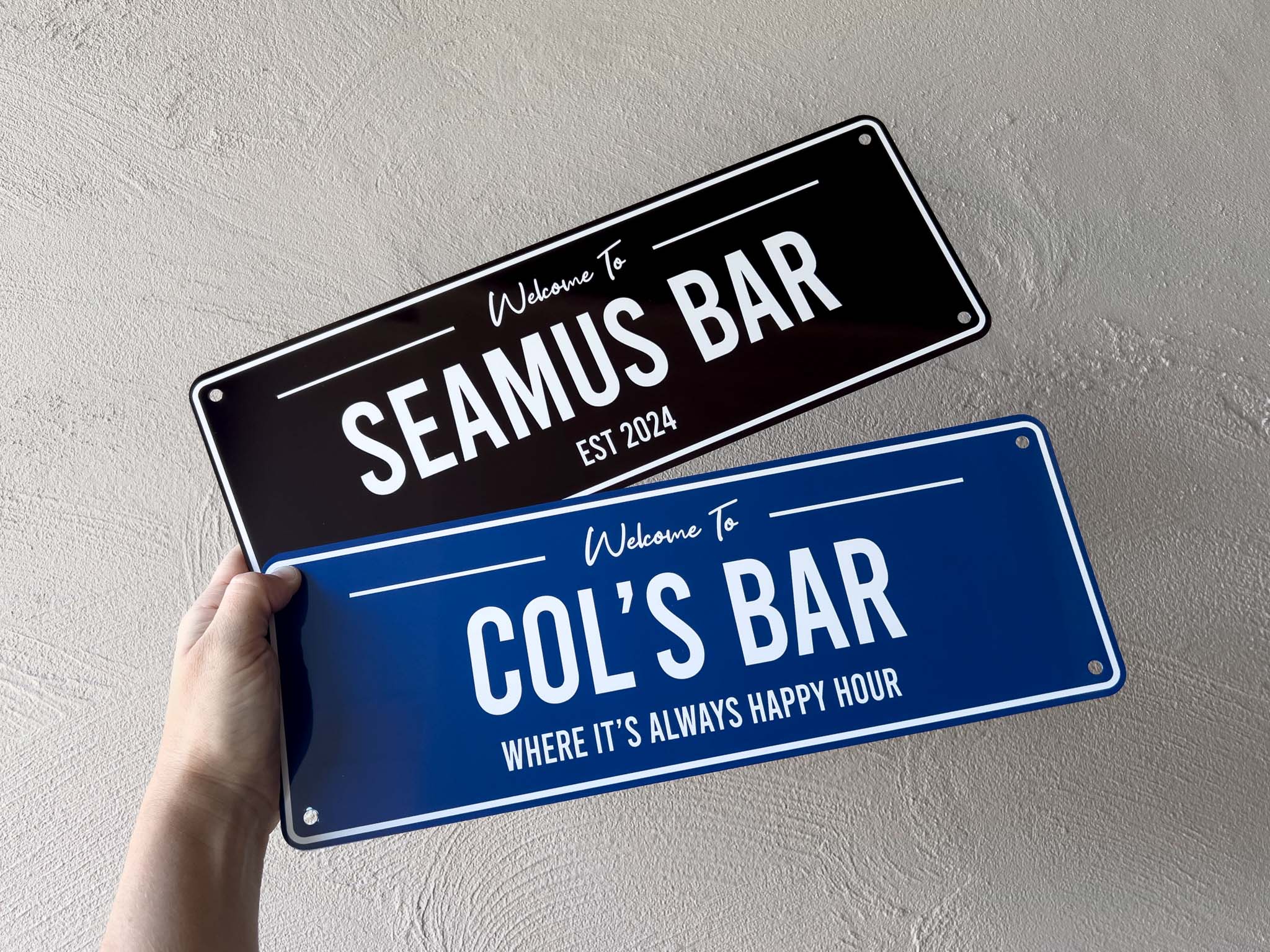 A black and white metal bar sign that says 'Seamus Bar' & a blue and white bar sign that says 'Col's Bar'.