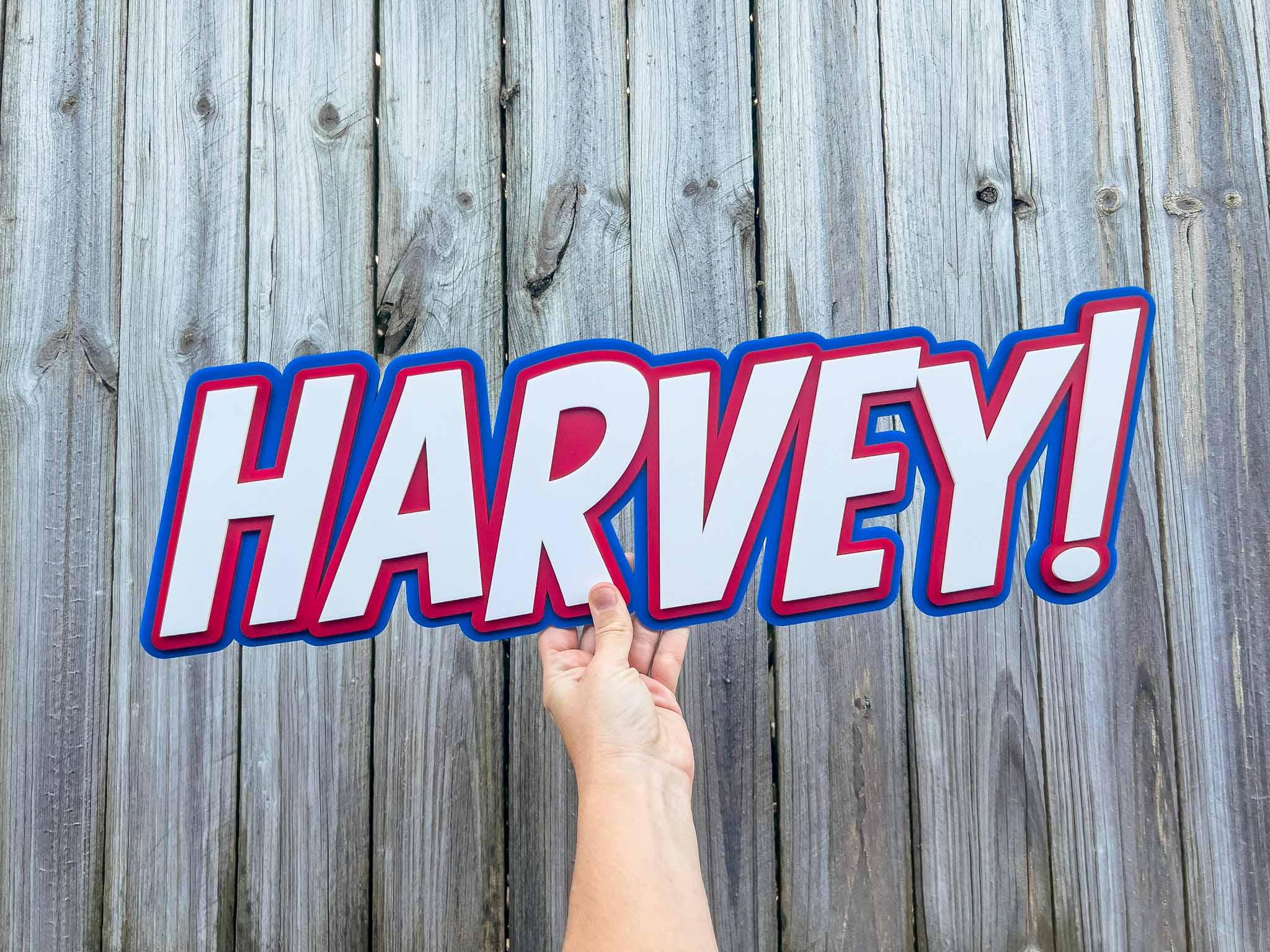 Hand holding a 3D acrylic layered name plaque sign with 'HARVEY!' in red and white against a wooden fence background