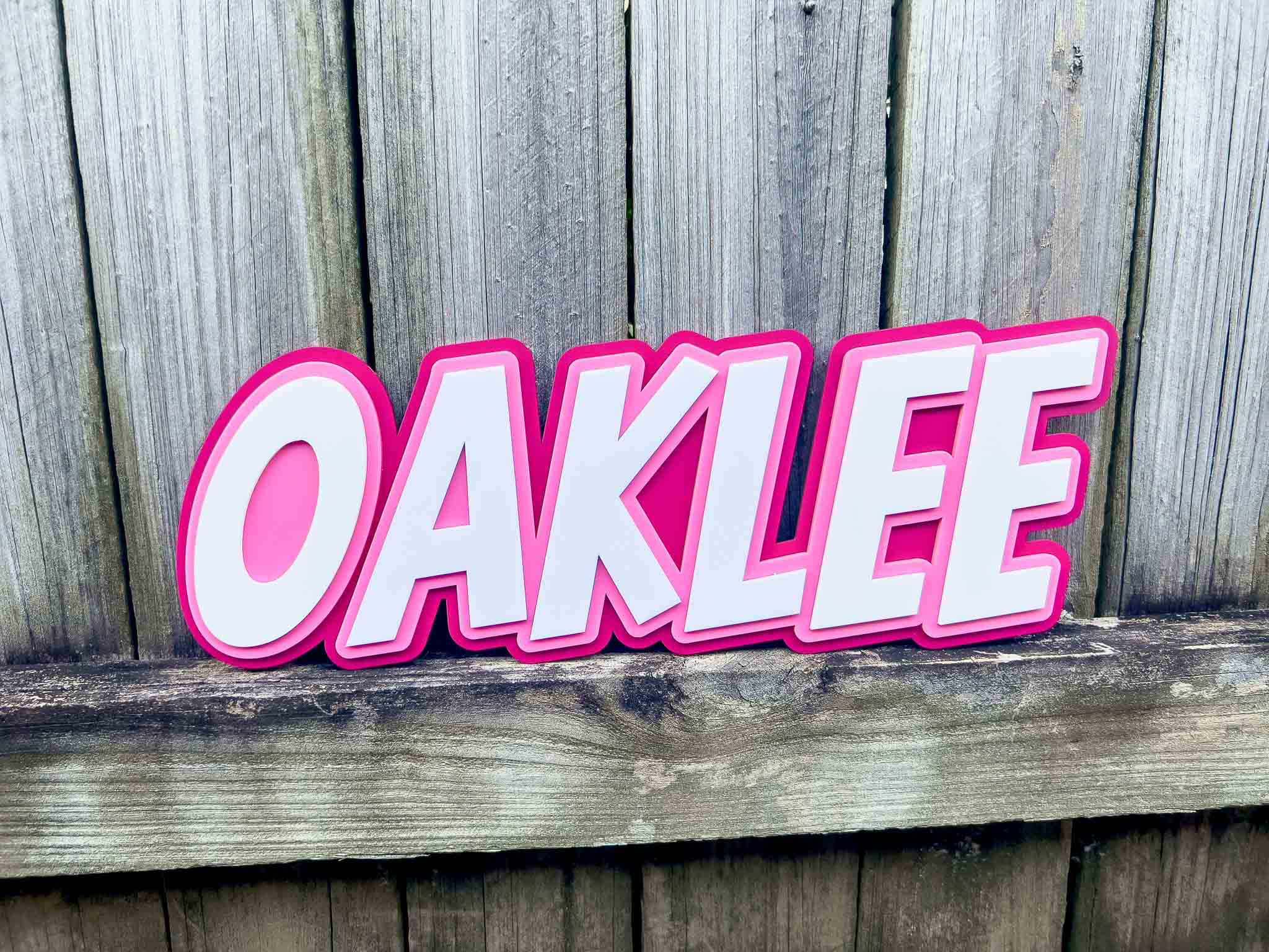 Pink and white 'OAKLEE' layered acrylic kid's name plaque displayed on a wooden fence background.