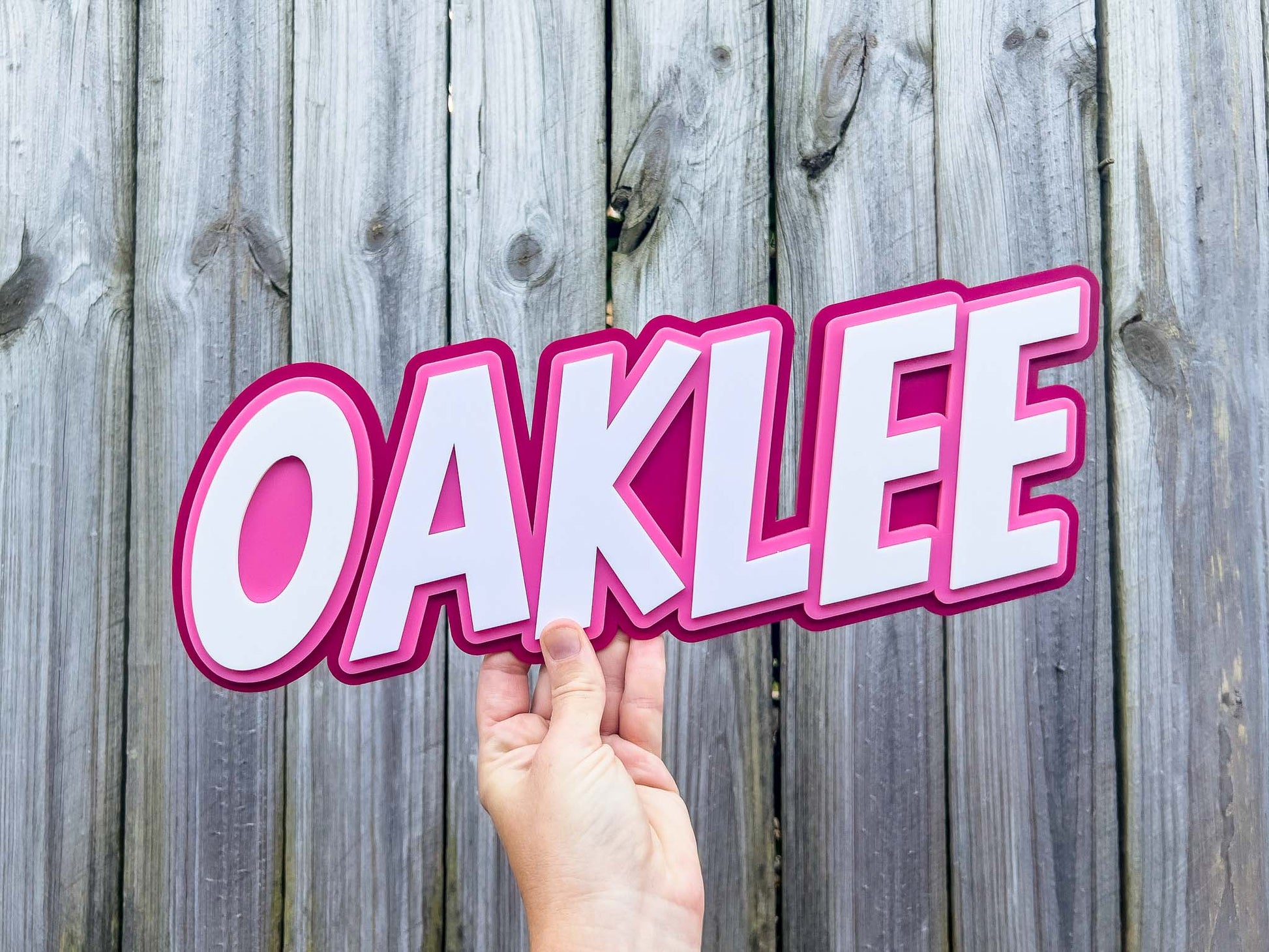 Hand holding a pink 'OAKLEE' sign against a wooden fence background