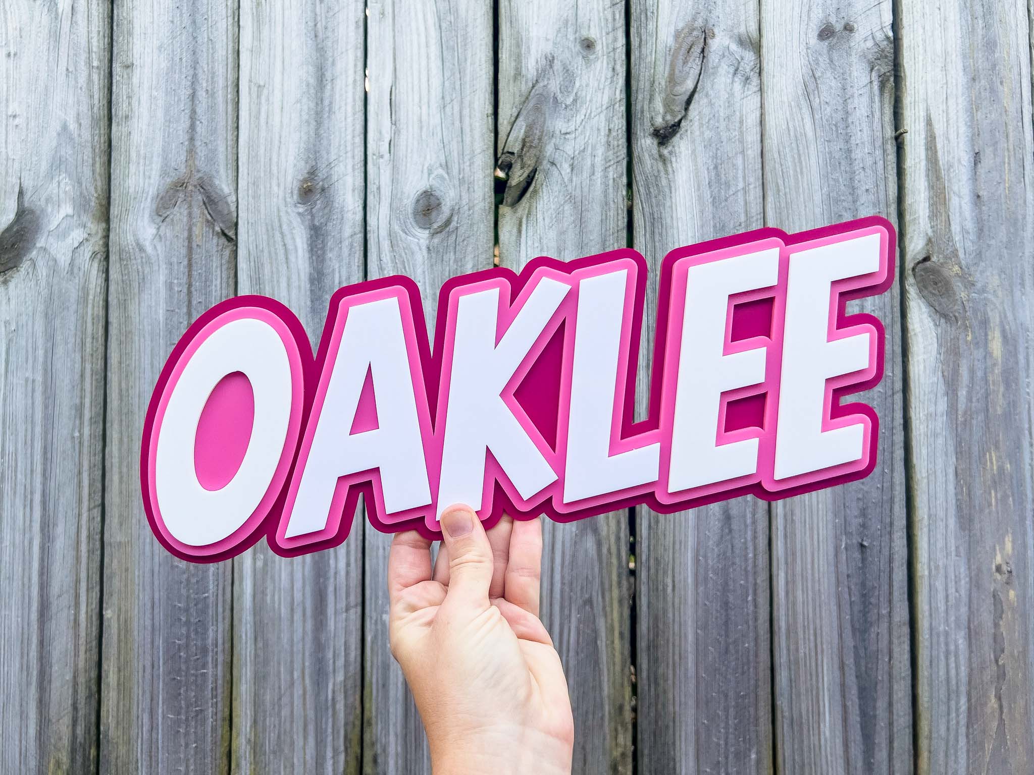 Hand holding a pink 'OAKLEE' sign against a wooden fence background