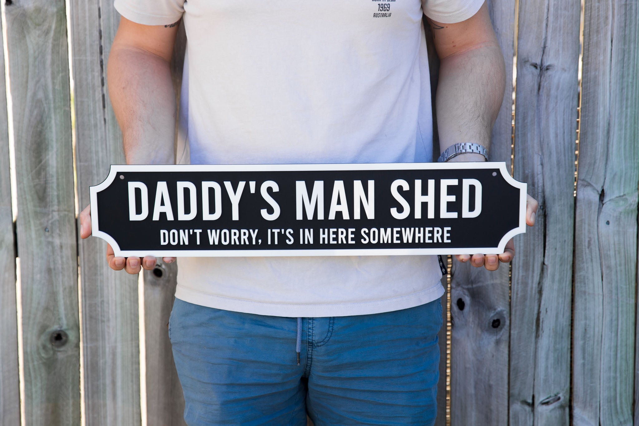 Person holding a sign that reads 'Daddy's Man Shed' against a wooden fence background.