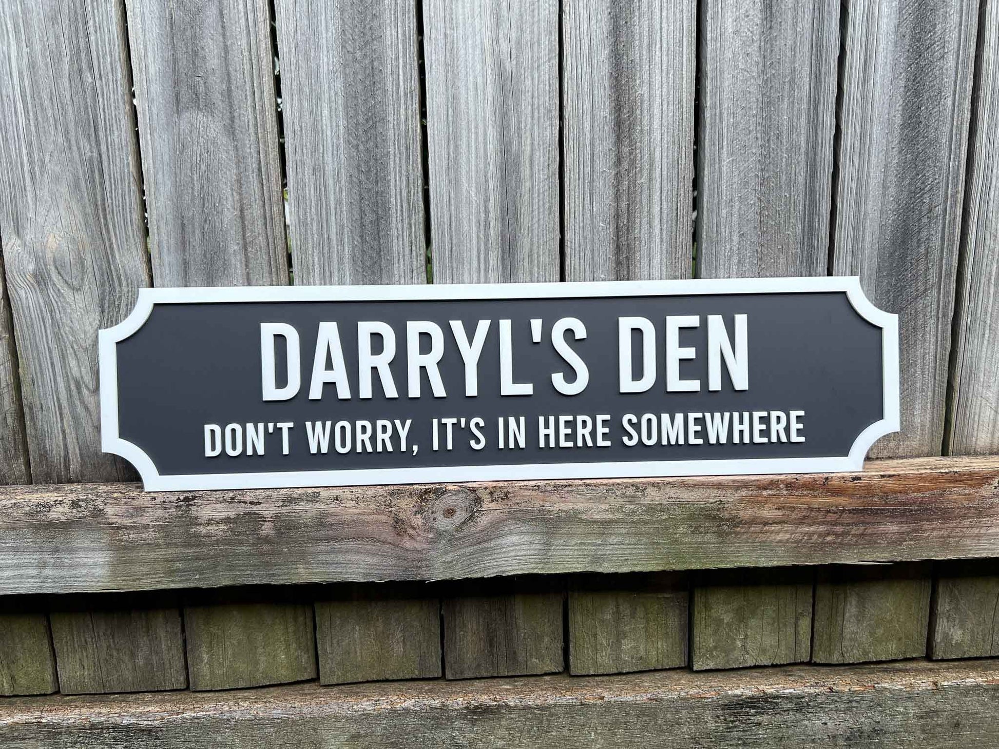 Decorative sign with 'Darryl's Den' text against a wooden fence background