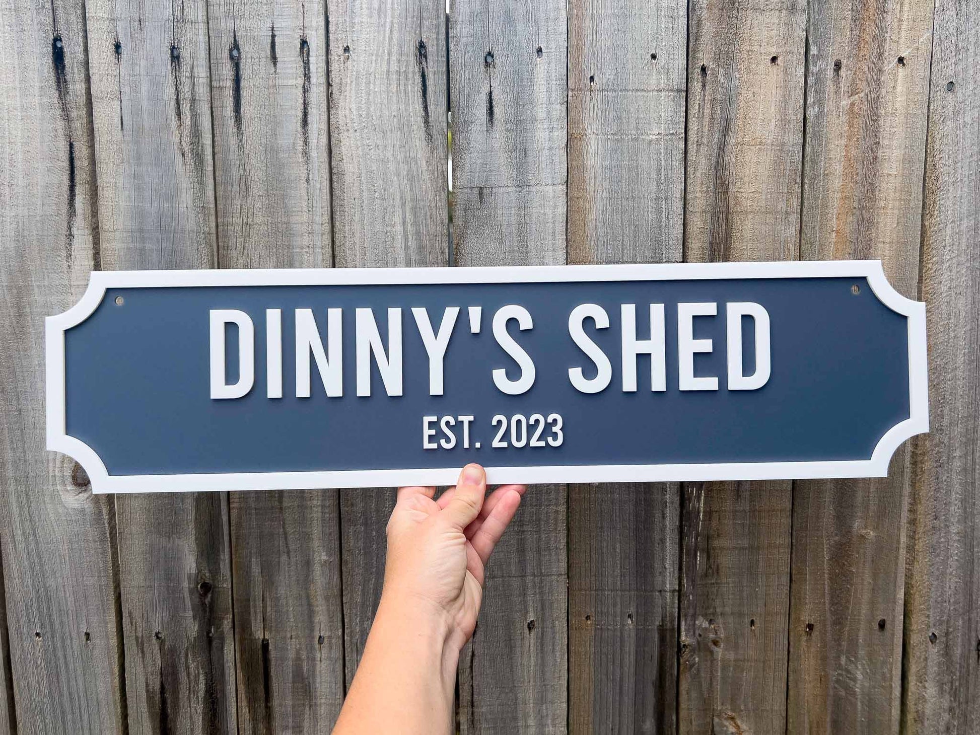 Classic vintage sign that reads 'DINNY'S SHED EST. 2023' against a wooden fence background.