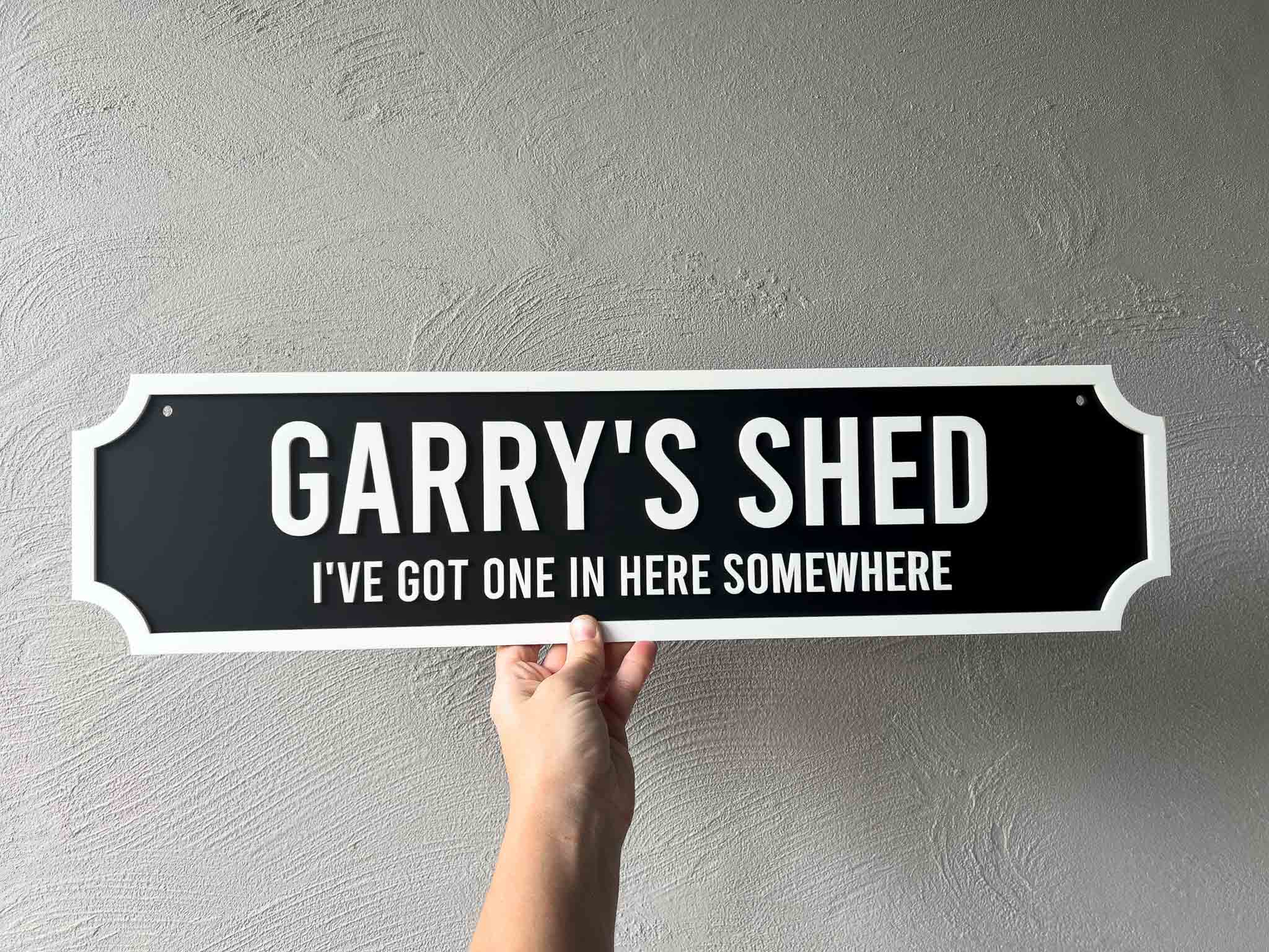 Hand holding a sign with 'GARRY'S SHED I'VE GOT ONE IN HERE SOMEWHERE' text against a gray wall.