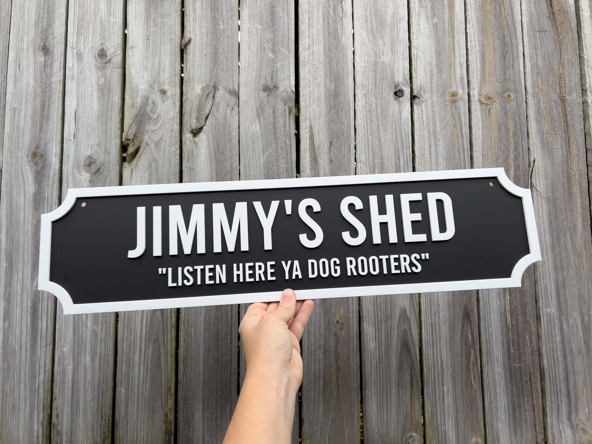 Hand holding a sign that reads 'Jimmy's Shed: Listen Here Ya Dog Rooters' against a wooden background.