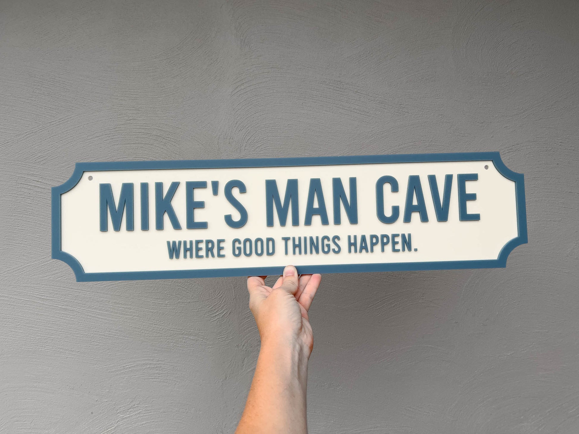 Hand holding a sign that reads 'MIKE'S MAN CAVE WHERE GOOD THINGS HAPPEN' in a cream and dusty blue colour.