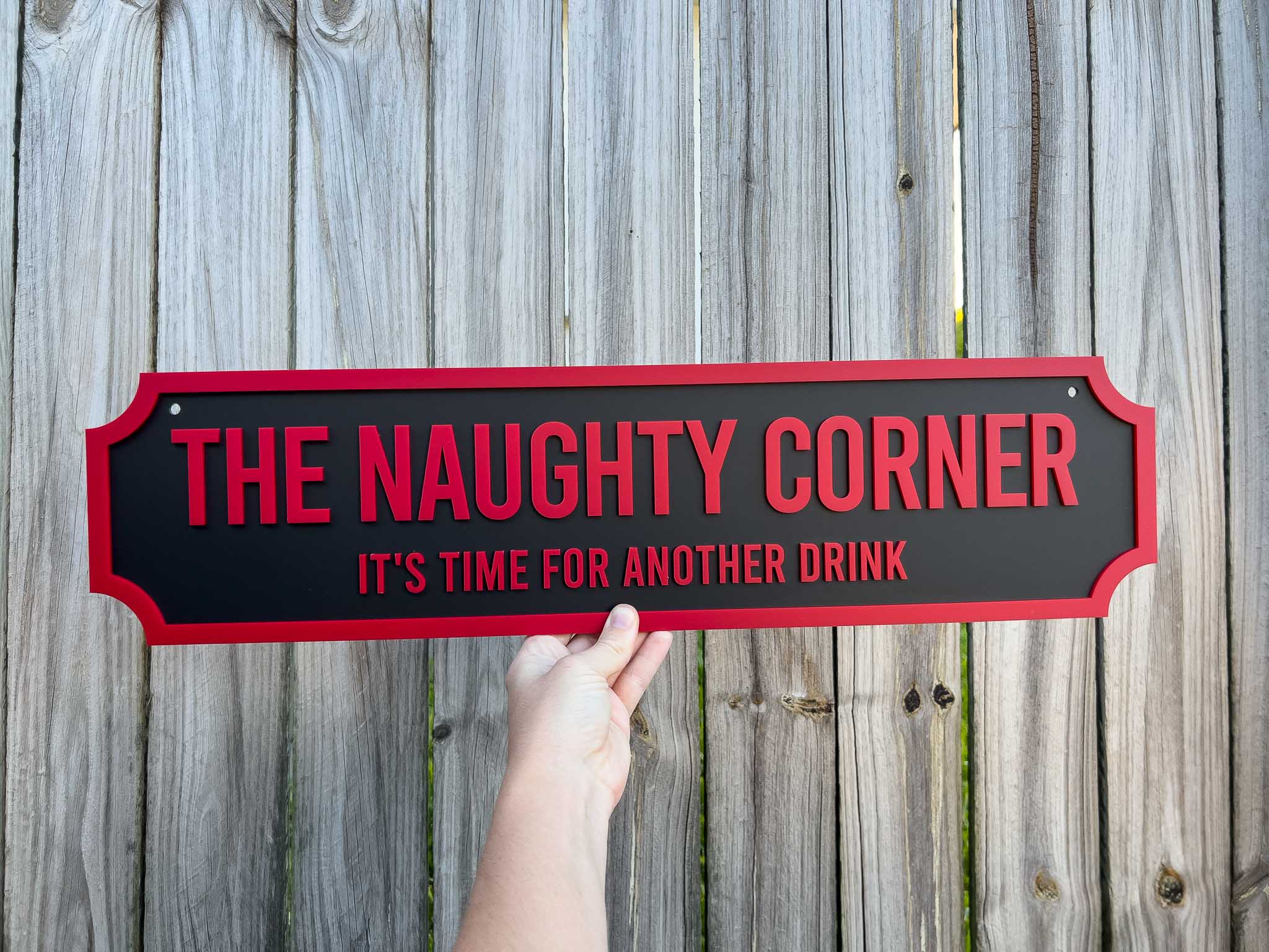 Hand holding a black and red 3D acrylic sign that reads 'The Naughty Corner. It's time for another drink' against a wooden background.