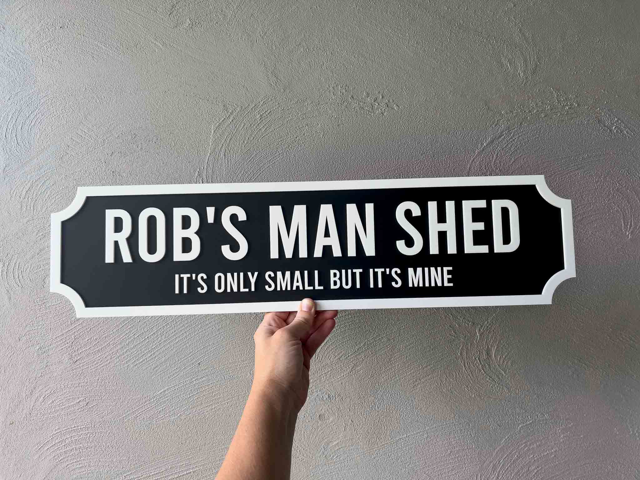 Hand holding a sign that reads 'Rob's Man Shed It's Only Small But It's Mine' against a gray wall.