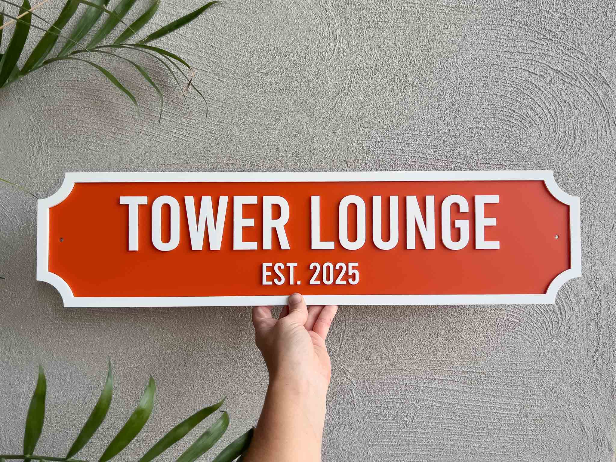 Handmade orange and white sign with 'TOWER LOUNGE EST. 2025' against a textured wall.