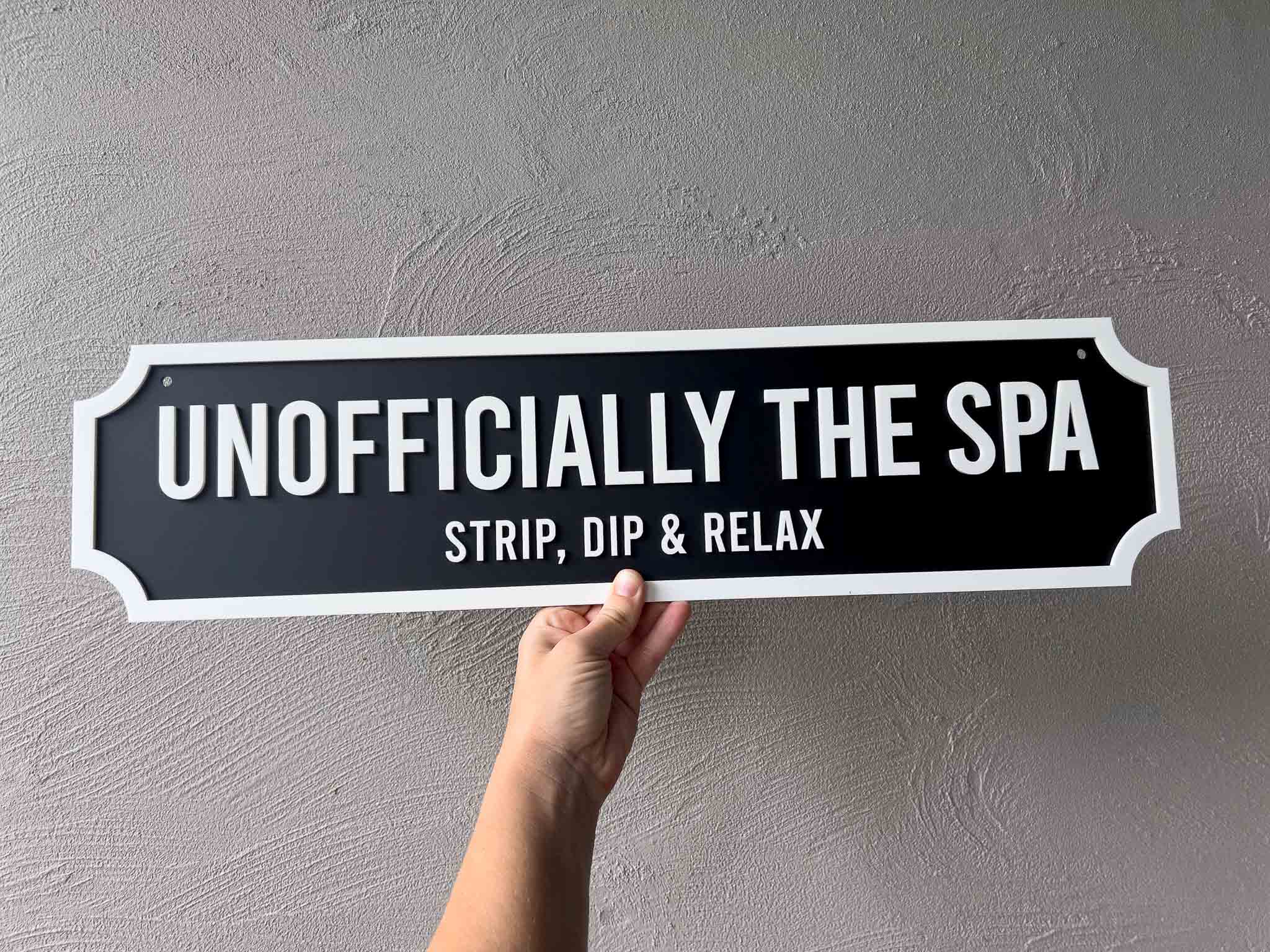 Hand holding a sign that reads 'Unofficially the Spa Strip, Dip & Relax' against a gray wall.