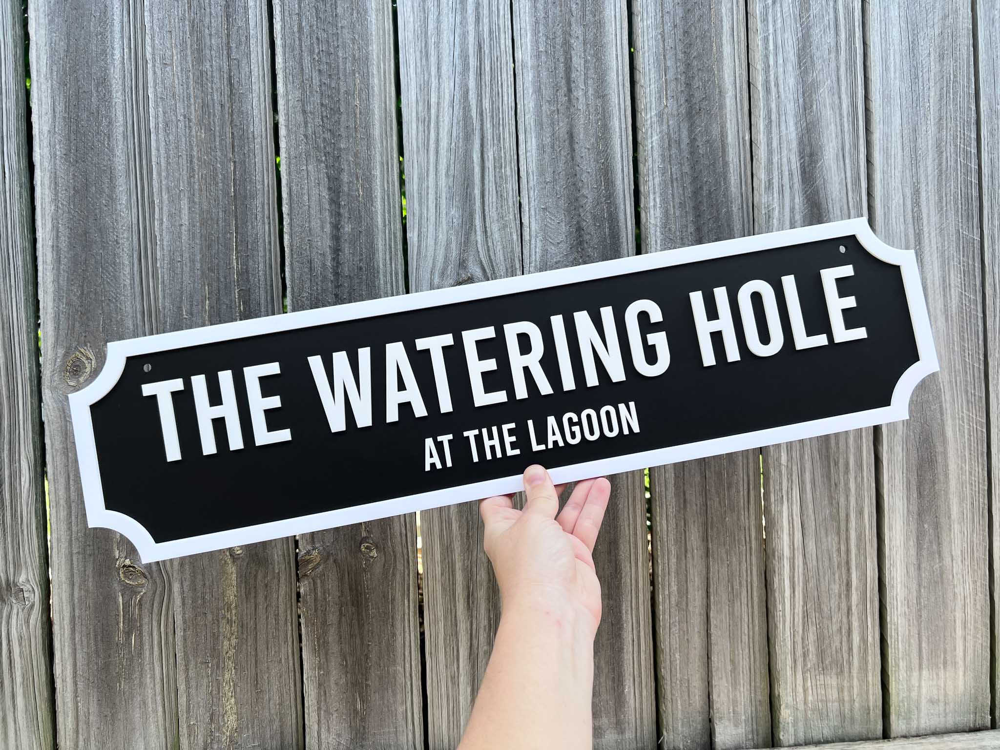 Hand holding a sign that reads 'The Watering Hole at the Lagoon' against a wooden fence background.