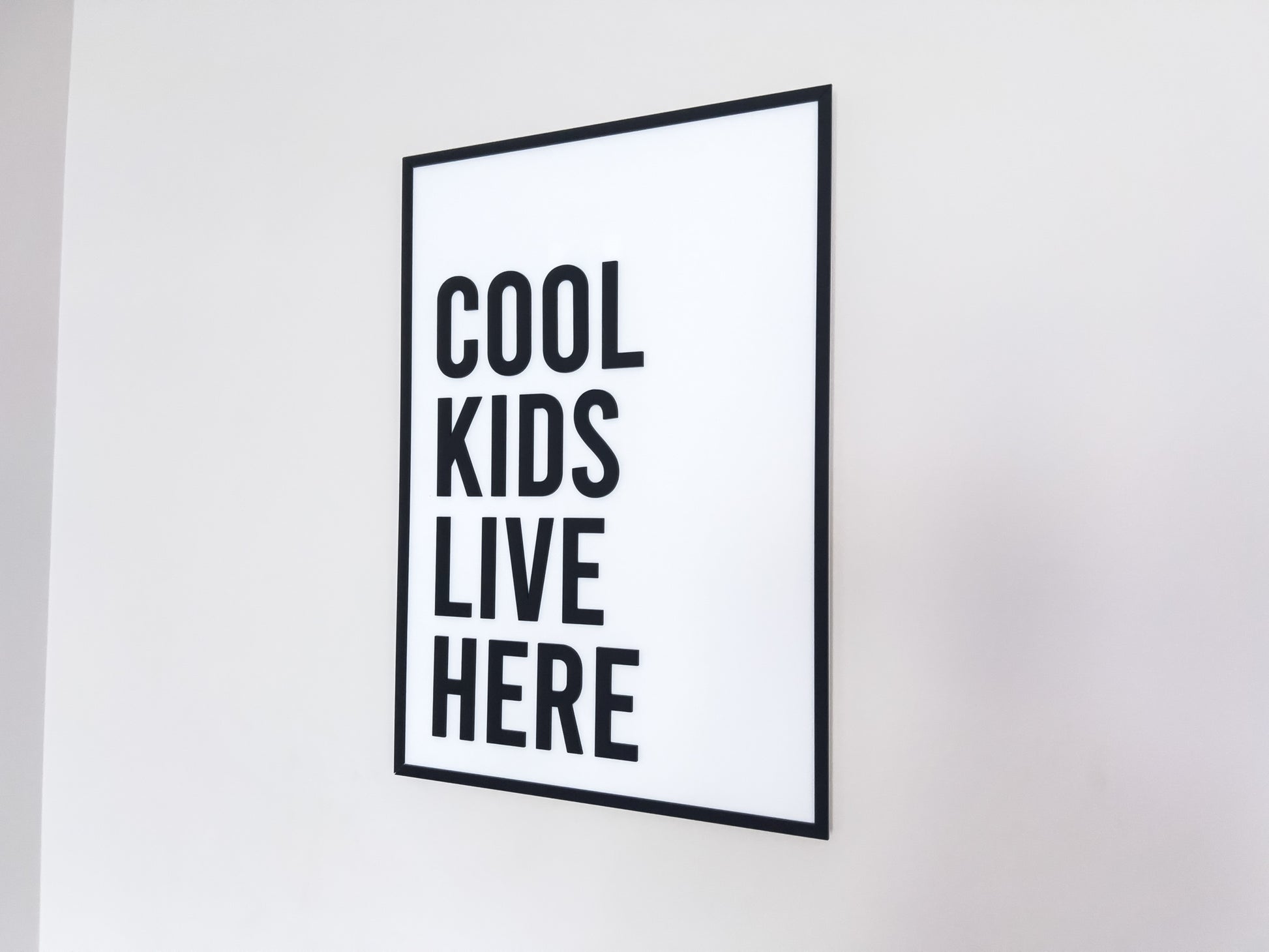 Framed black and white 3D acrylic sign with 'Cool kids live here' text hanging on a white wall.