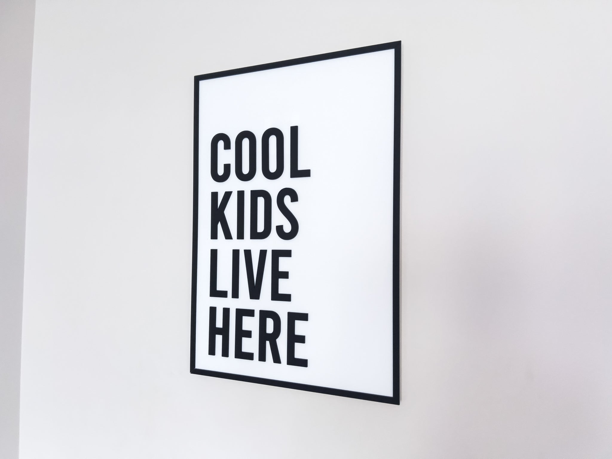Framed black and white 3D acrylic sign with 'Cool kids live here' text hanging on a white wall.