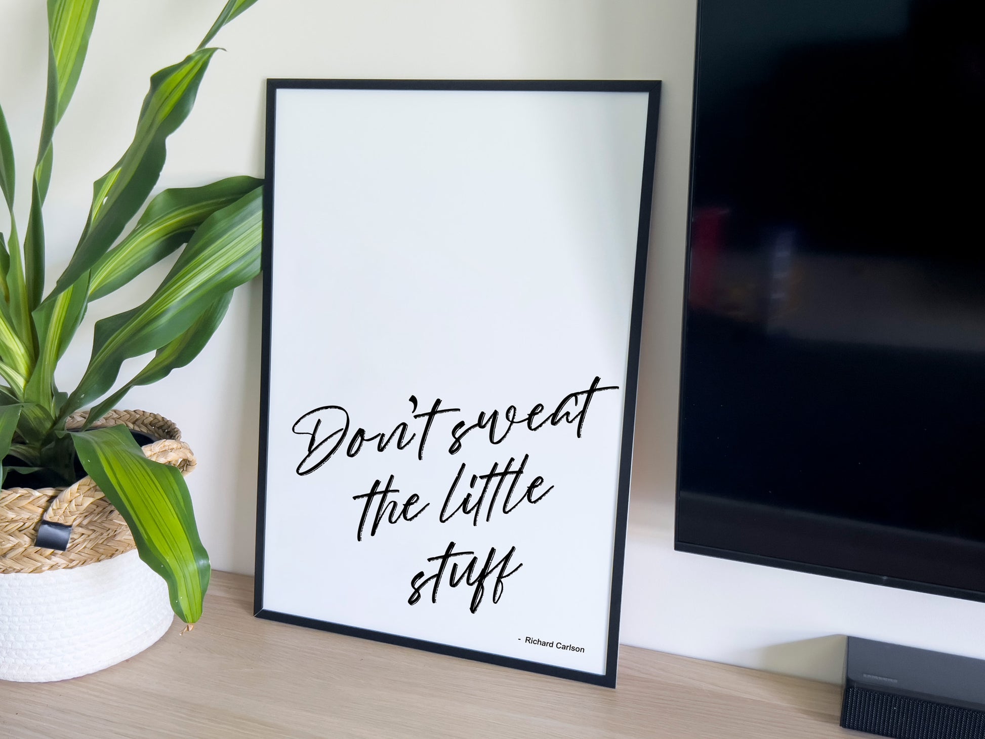Framed print with motivational quote 'Don't sweat the little stuff' on a desk with a plant and TV in the background.