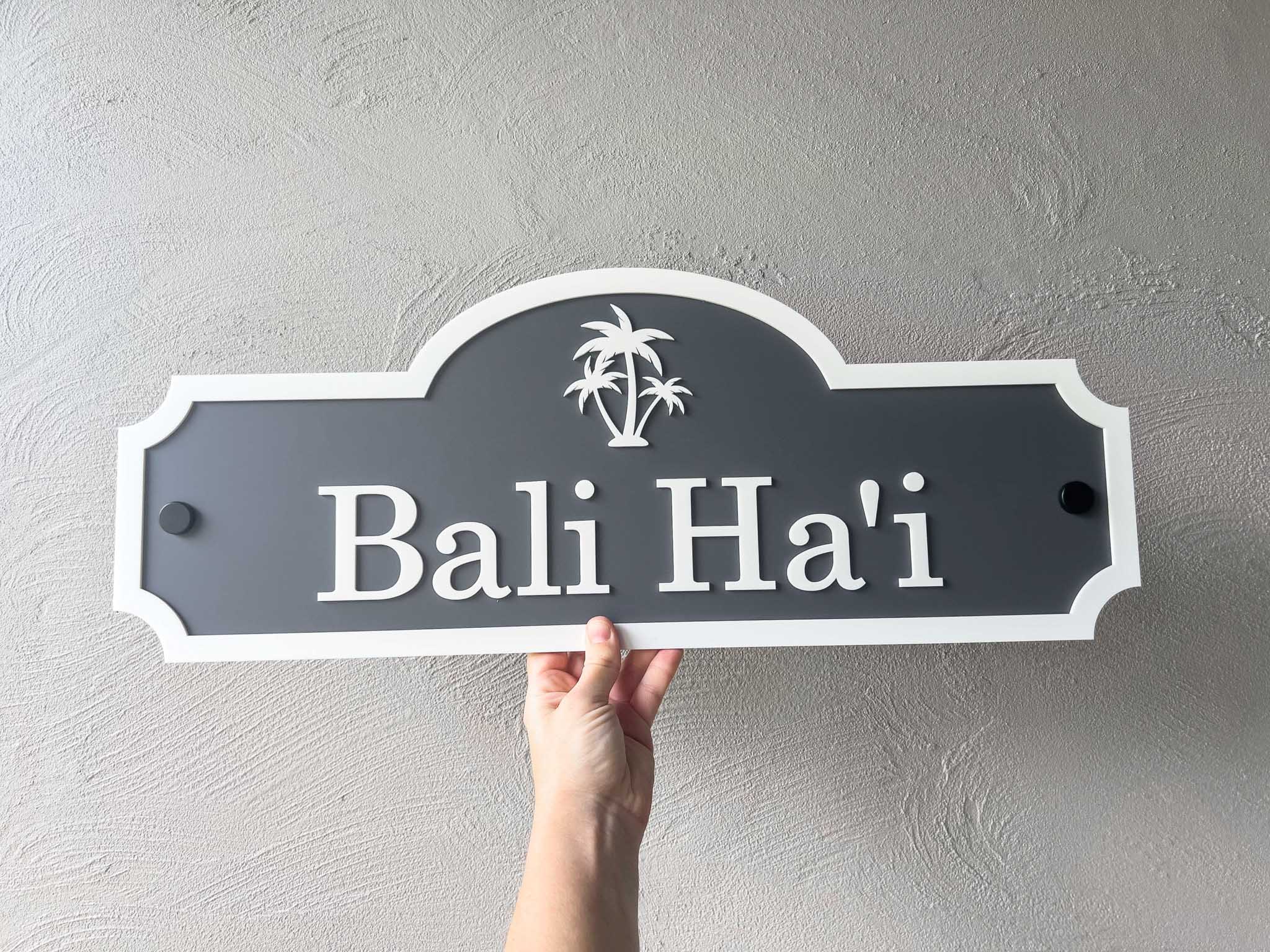 Hand holding a custom made sign in dark grey and white with 'Bali Ha'i' text against a plain background