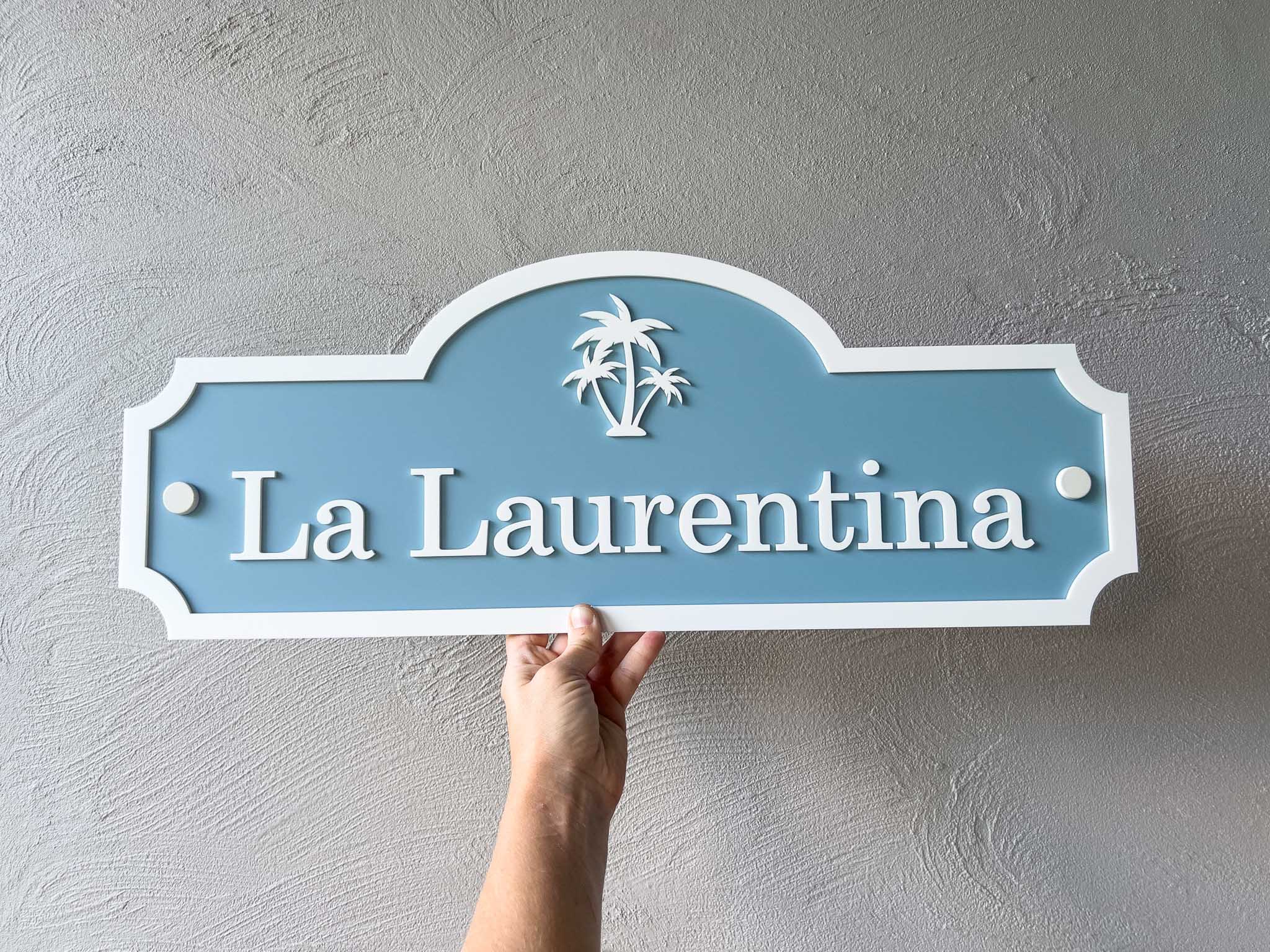 Hand holding a decorative coastal blue and white vintage palm tree style sign that says 'La Laurentina'.
