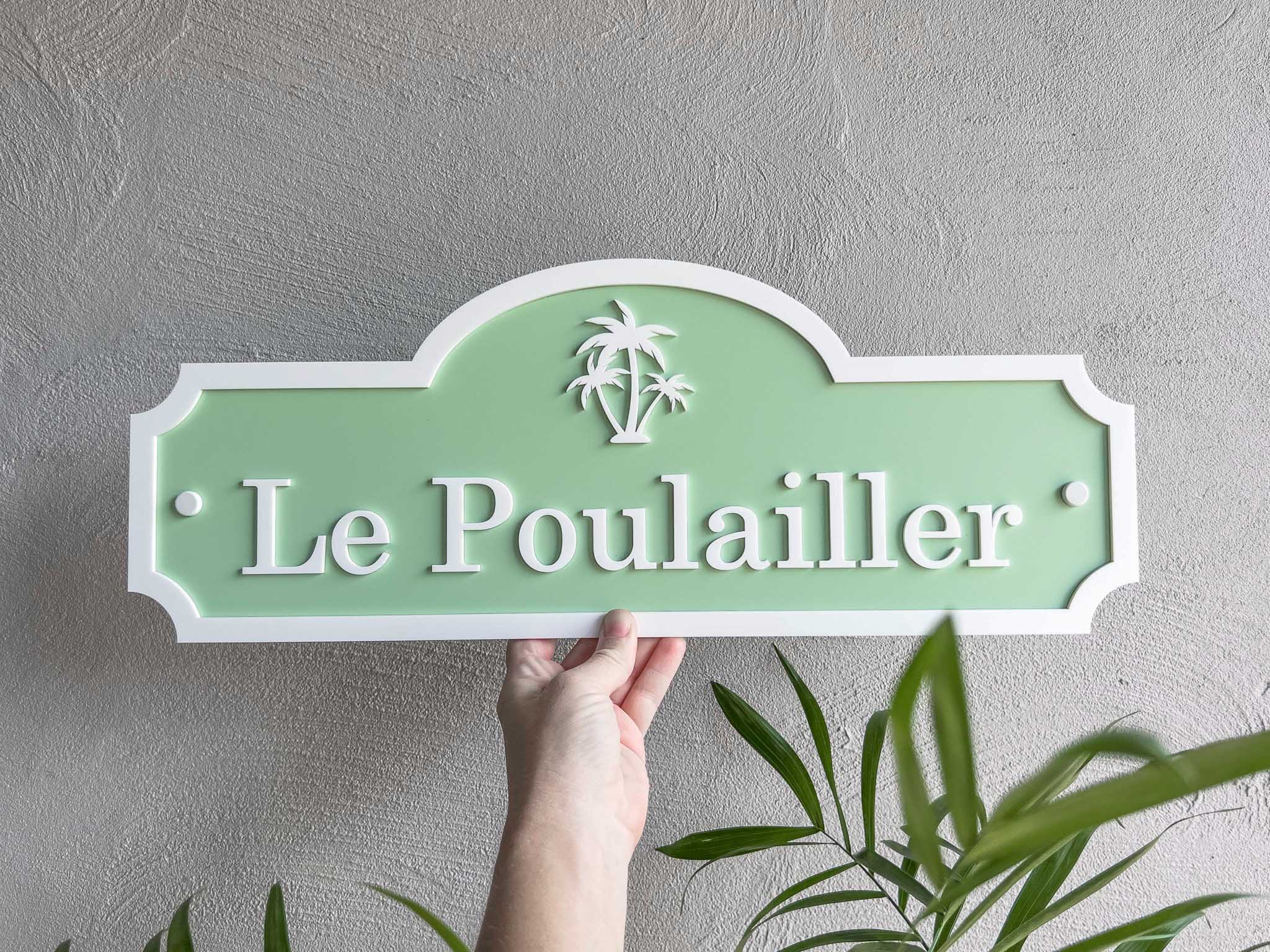 Hand holding a tropical green sign with 'Le Poulailler' against a gray wall.