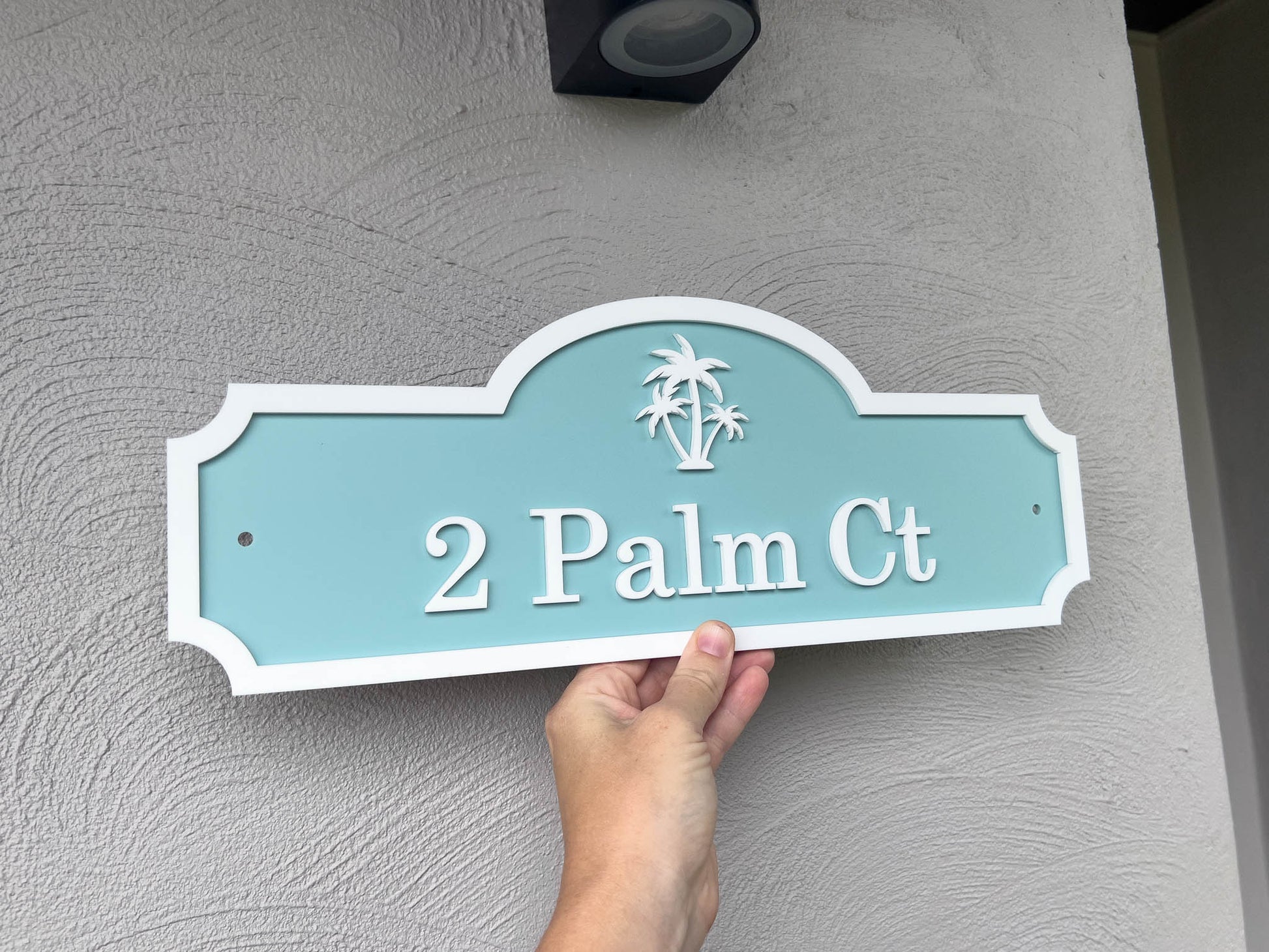 Hand holding a turquoise house sign with 'Palm Ct' on a gray wall.