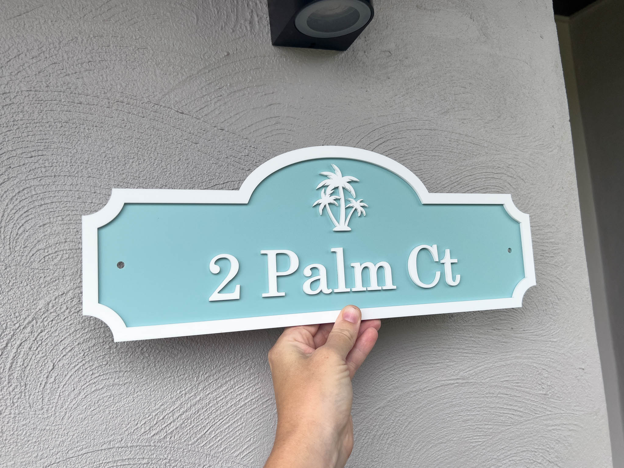 Hand holding a turquoise house sign with 'Palm Ct' on a gray wall.