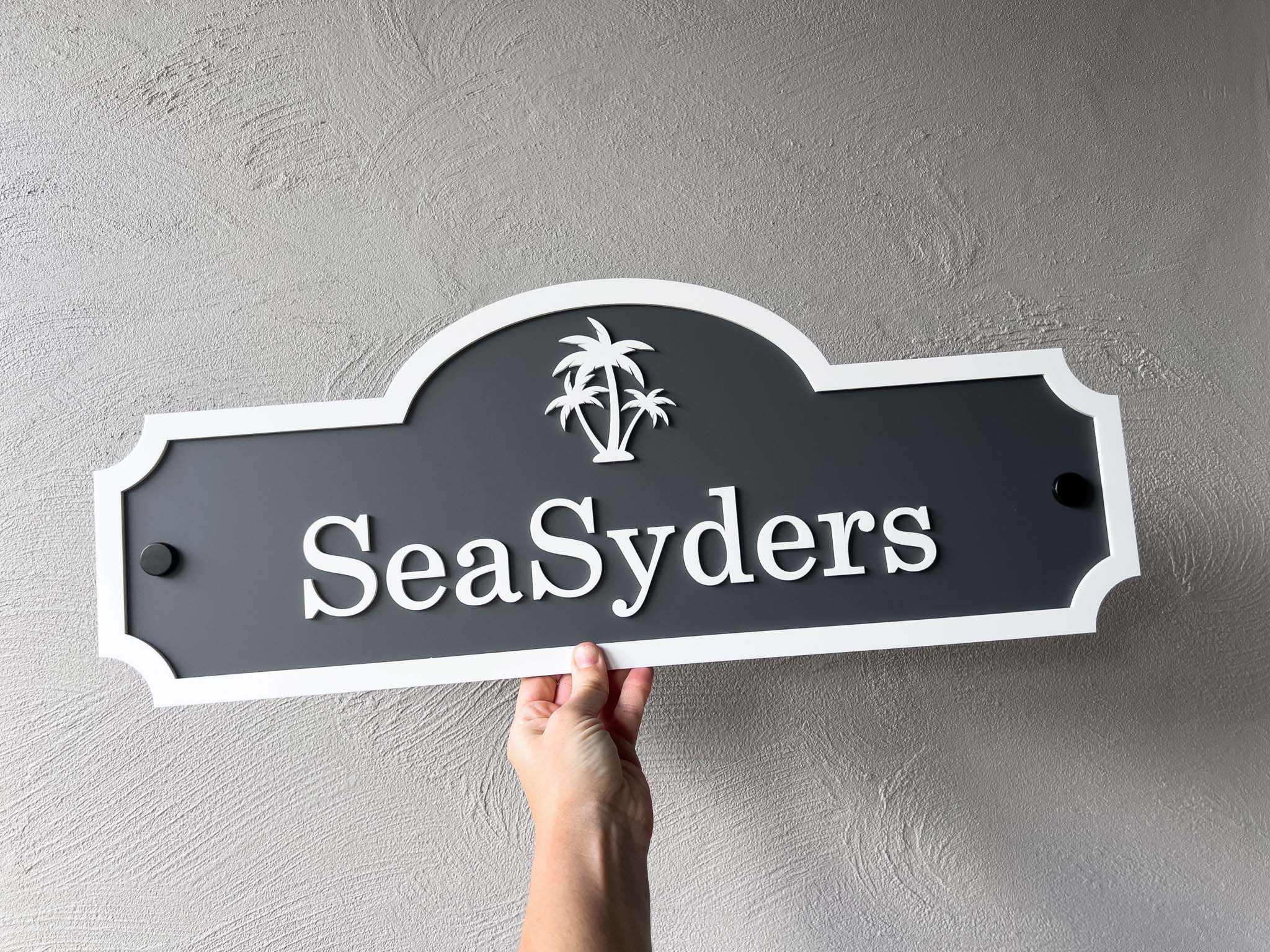 Hand holding a custom made 3D acrylic sign with 'SeaSyders' and palm tree design against a grey background