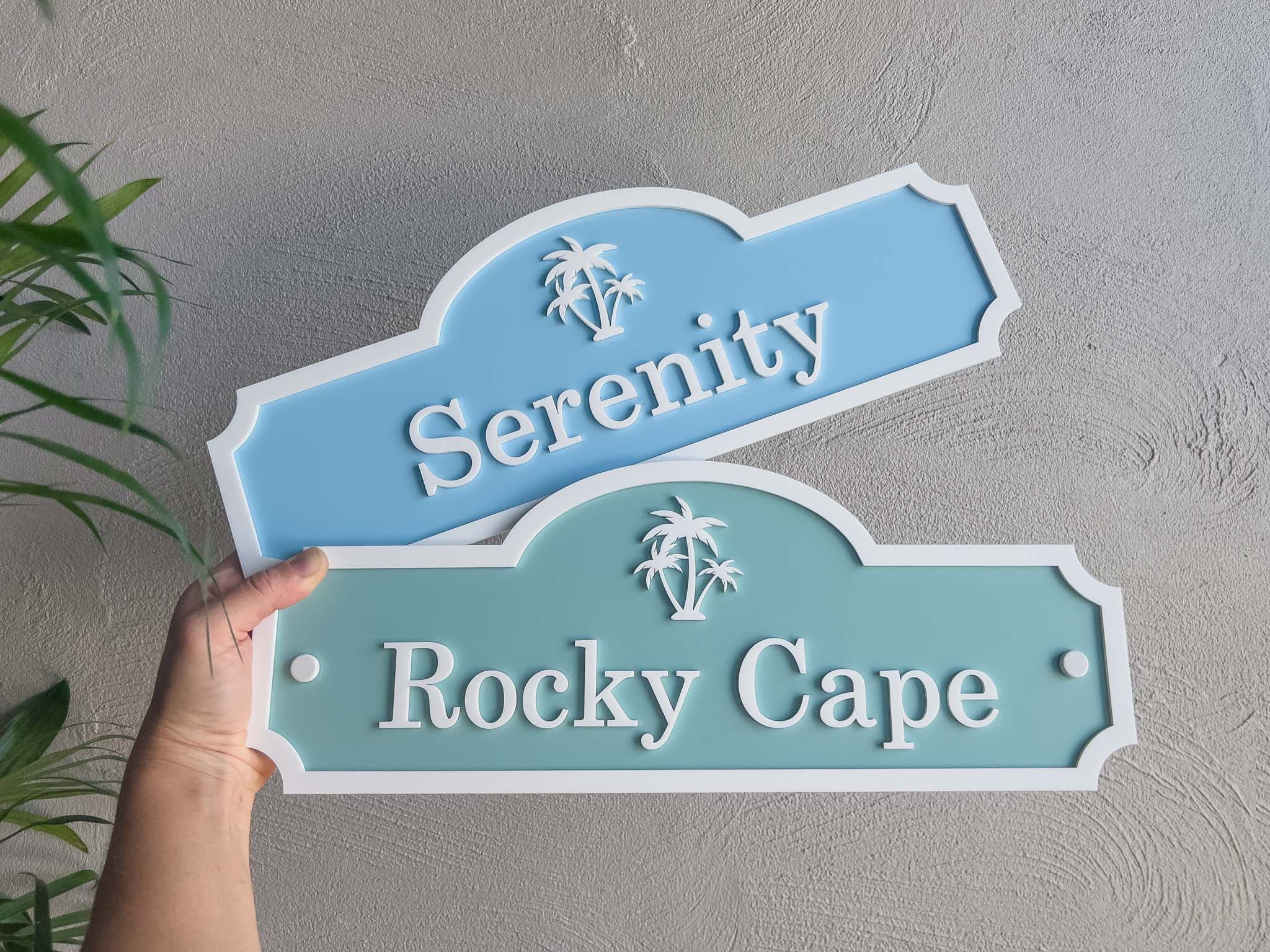 Two decorative palm tree signs with 'Serenity' and 'Rocky Cape' text on a gray background.