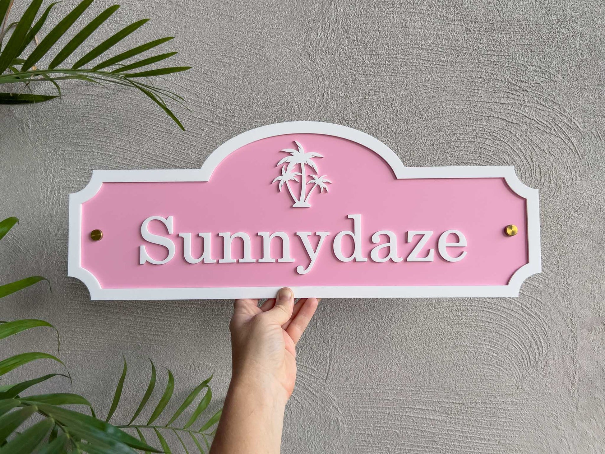 Hand holding a tropical pink and white sign with 'Sunnydaze' on a gray wall with green plants.